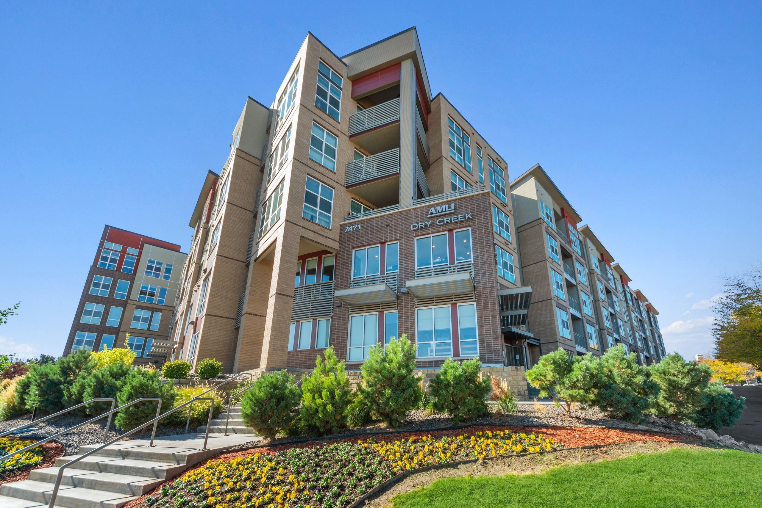 Daytime exterior of AMLI Dry Creek apartment building with brick façade and landscaping with flowers and bushes and blue sky