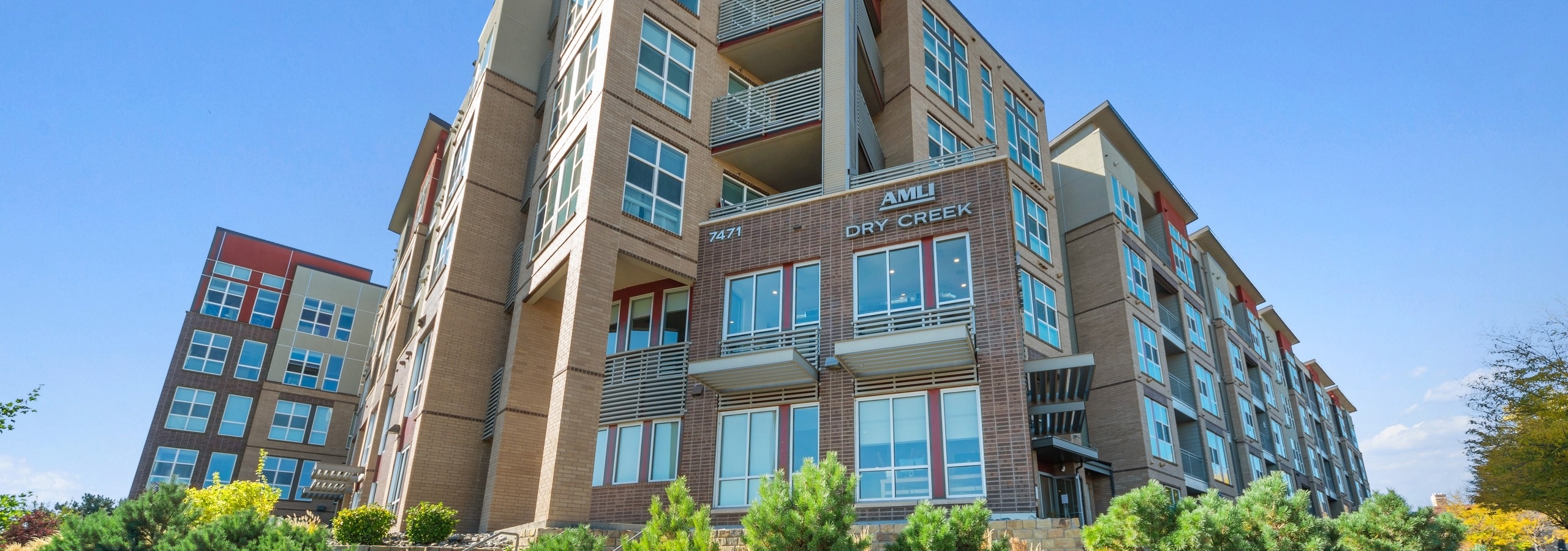 Daytime exterior of AMLI Dry Creek apartment building with brick façade and landscaping with flowers and bushes and blue sky