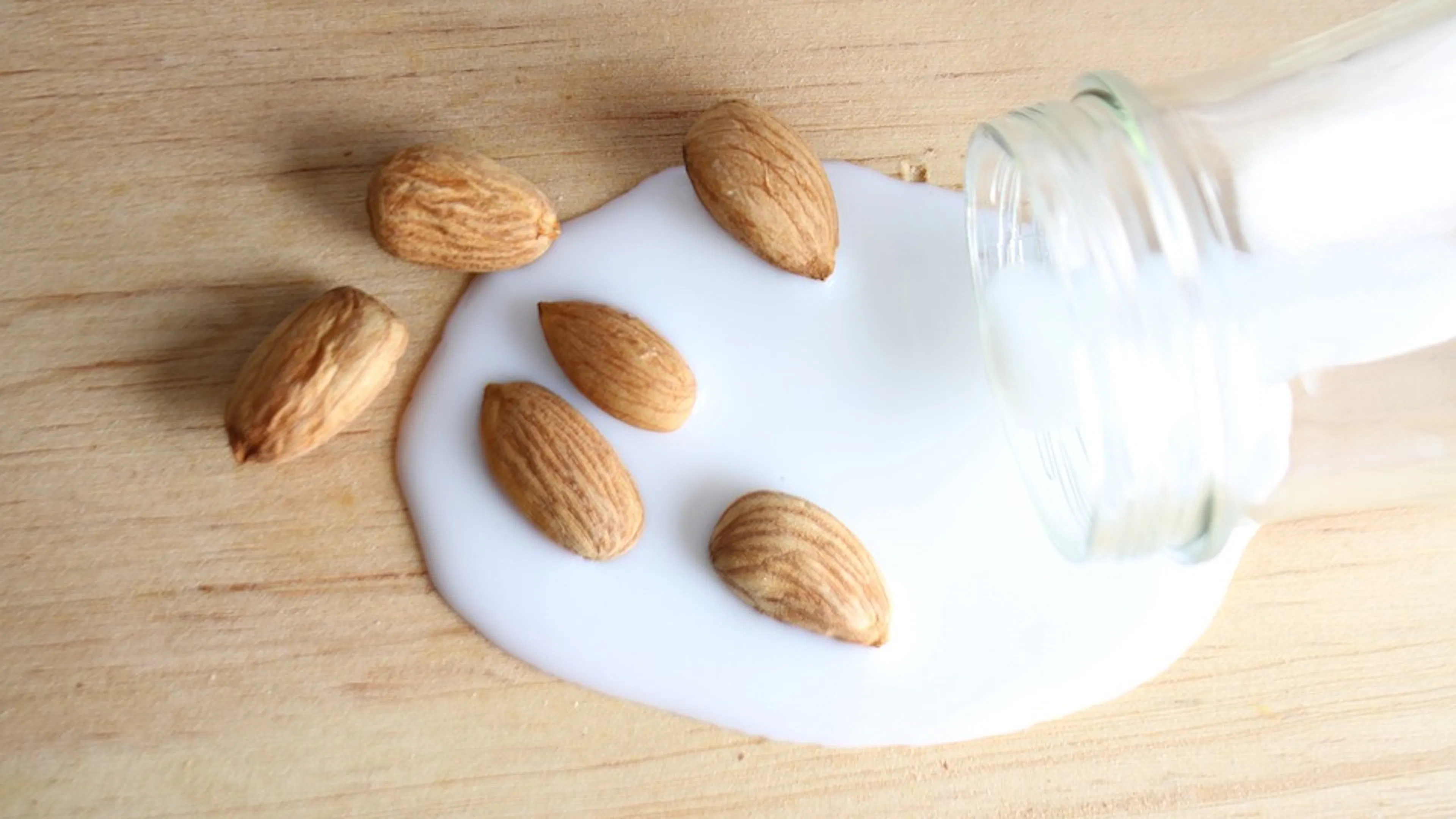 A glass jar toppled over on a wooden table with milk spilling out of it and whole almonds sprinkled on the milk