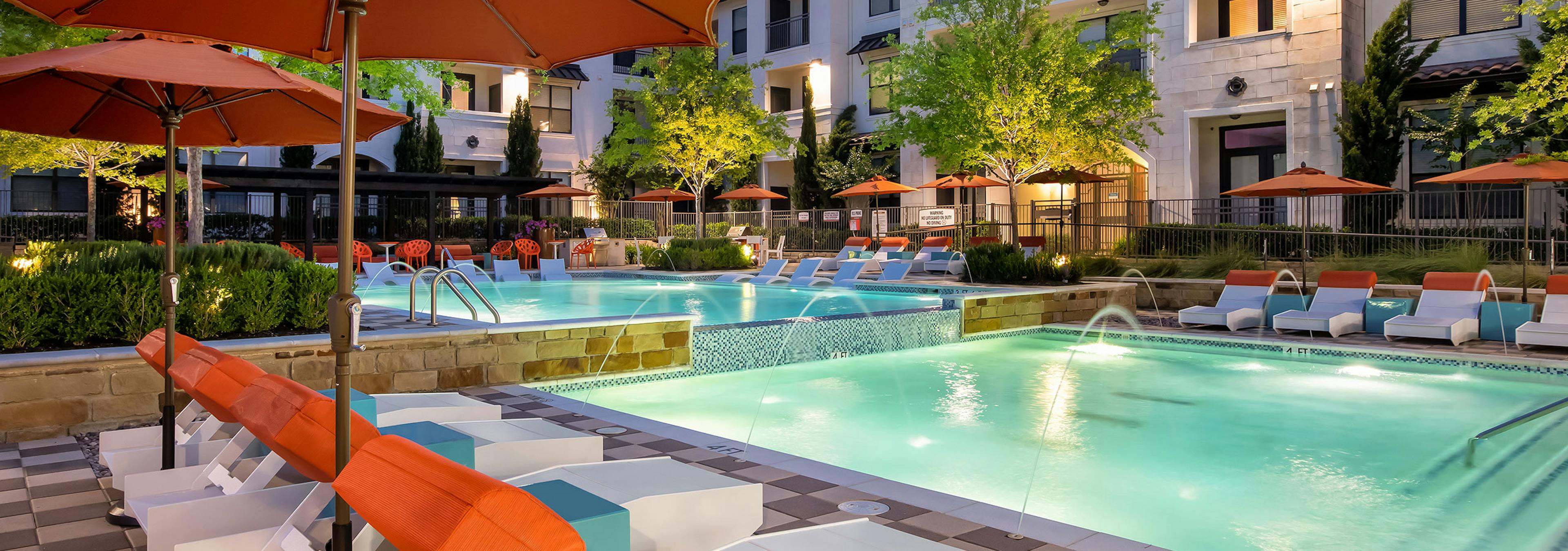 Pool at AMLI on Riverside at night with orange umbrellas, white lounge chairs and vibrant trees surrounding the water