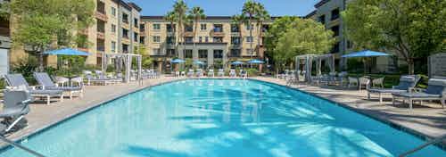 Daytime view of large pool at AMLI Warner Center apartments with blue cushioned lounge chairs and cabanas and umbrellas