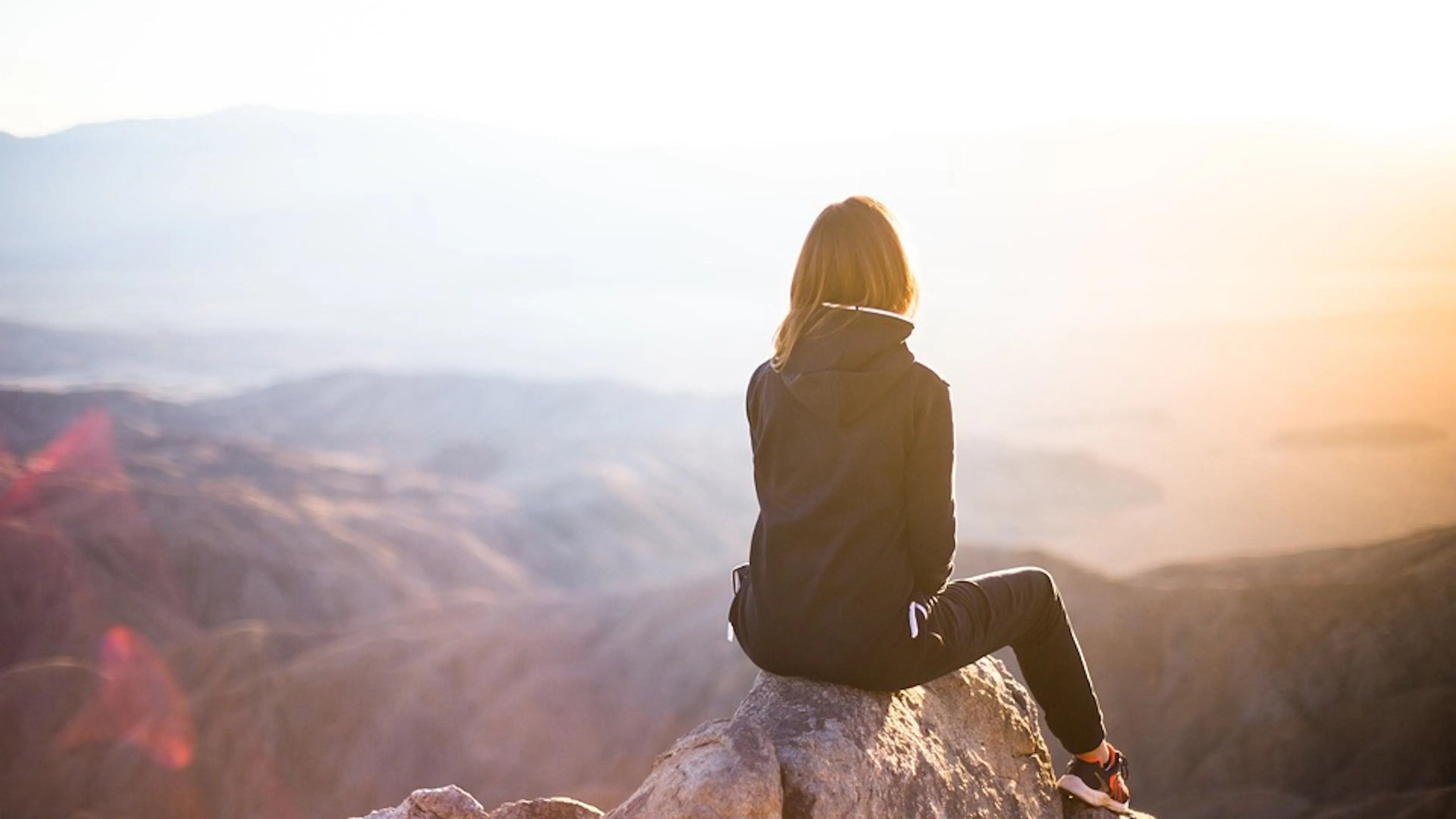 A woman sitting on a rocky mountain peak overlooking a mountain range and a lake during sunset