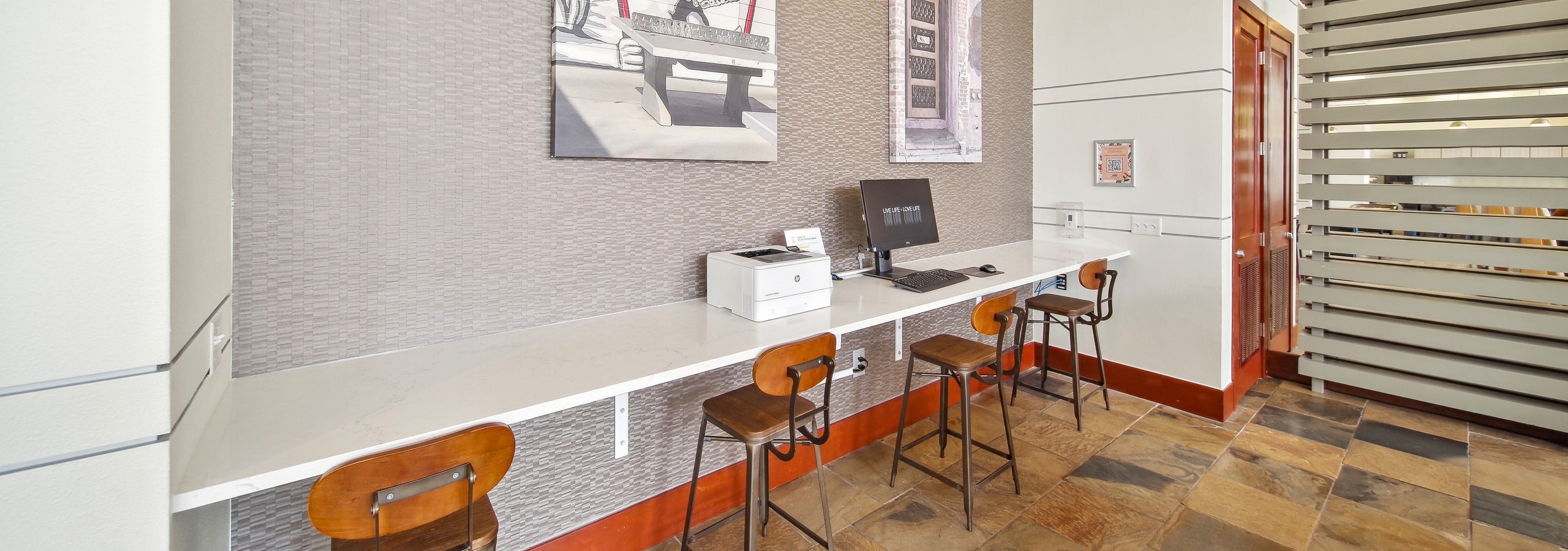 A business center at AMLI Park Avenue featuring a computer and printer with high top wooden chairs and two canvas art pieces