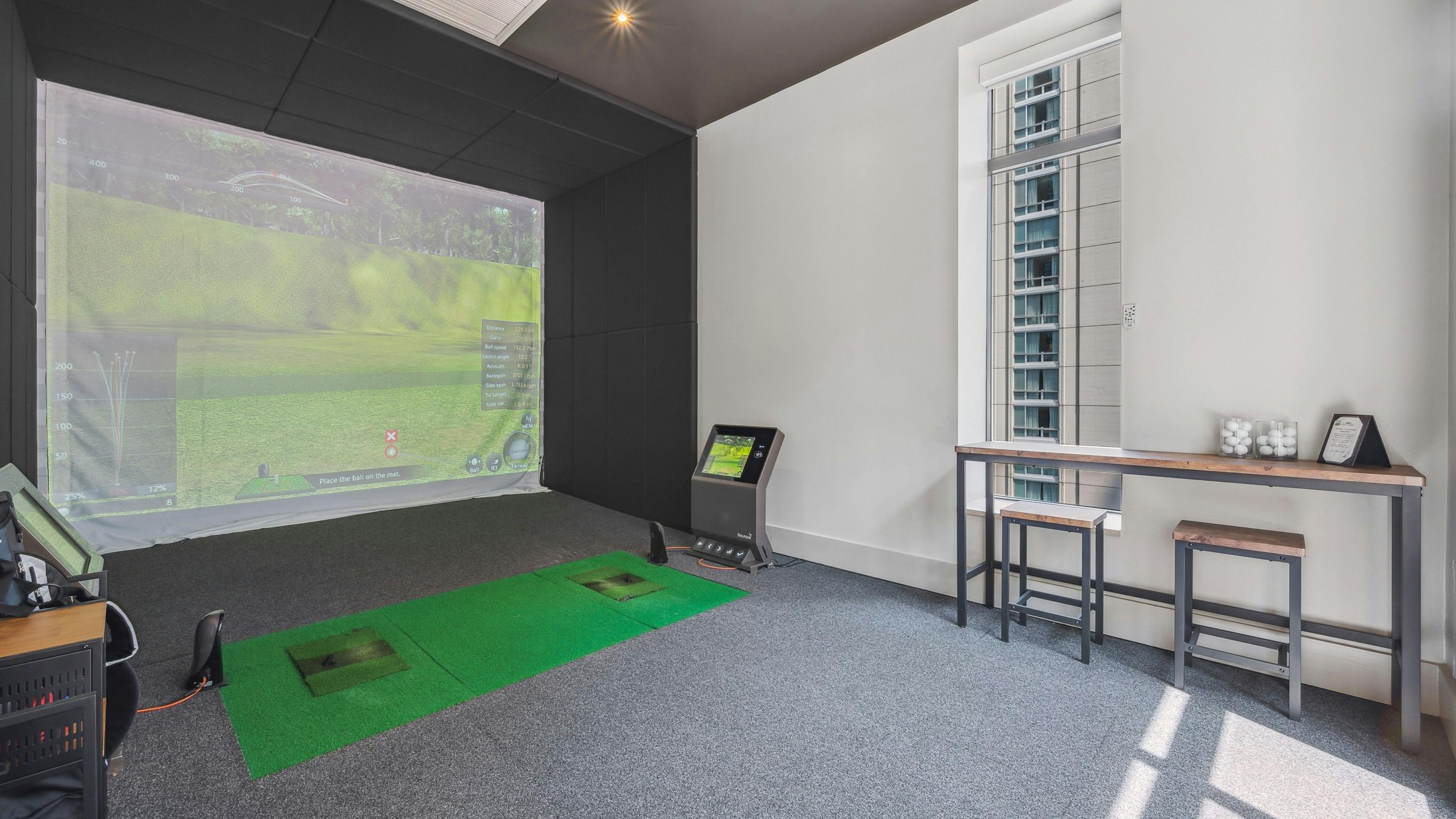 Golf simulator at AMLI River North with large screen with golf scene and a wood table with 2 stools with gray carpeting 