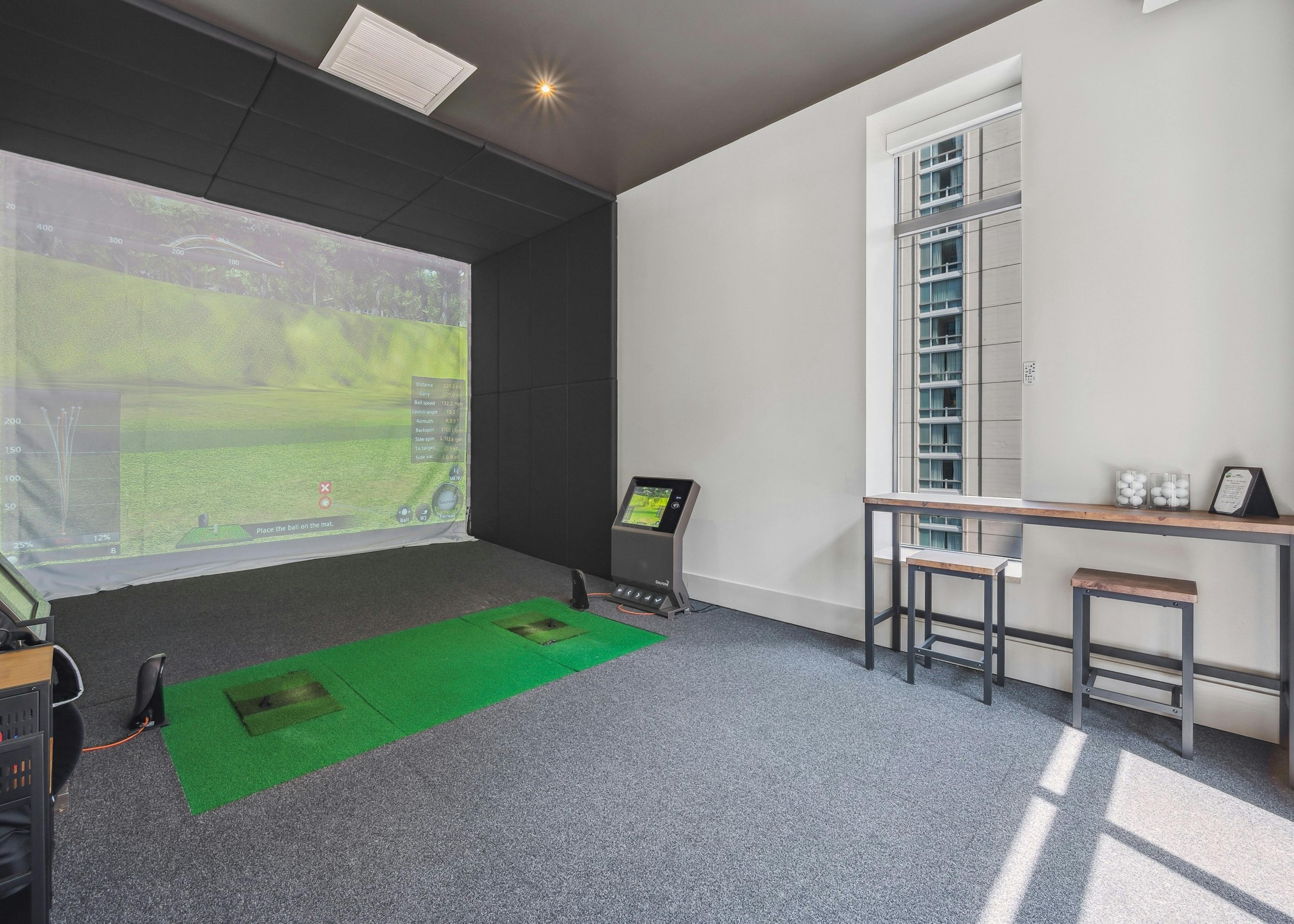 Golf simulator at AMLI River North with large screen with golf scene and a wood table with 2 stools with gray carpeting 