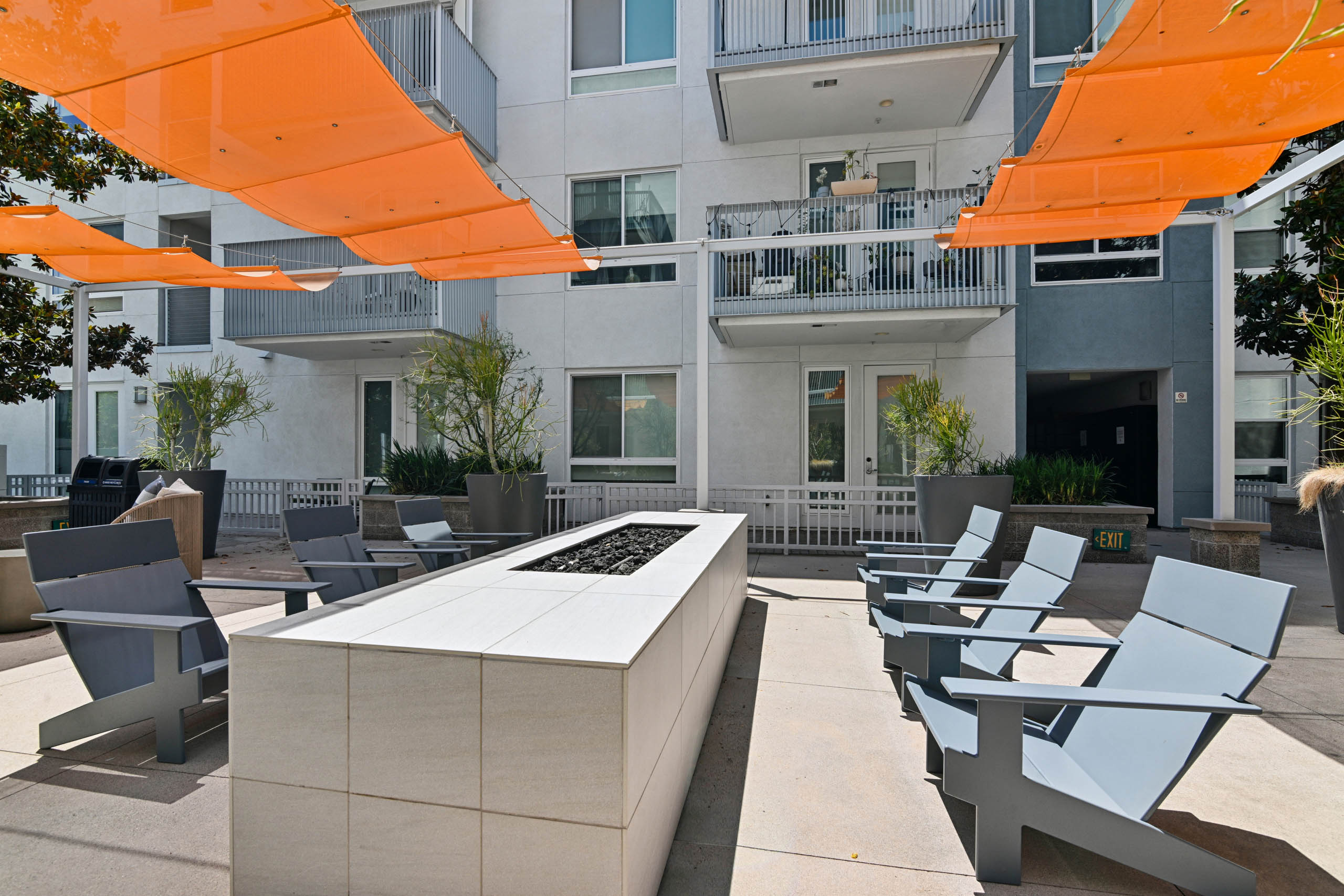 Courtyard at AMLI Lex on Orange with a long fire pit surrounded by gray lounge chairs, orange shade sails overhead, and large potted plants, set against the building’s balconies.