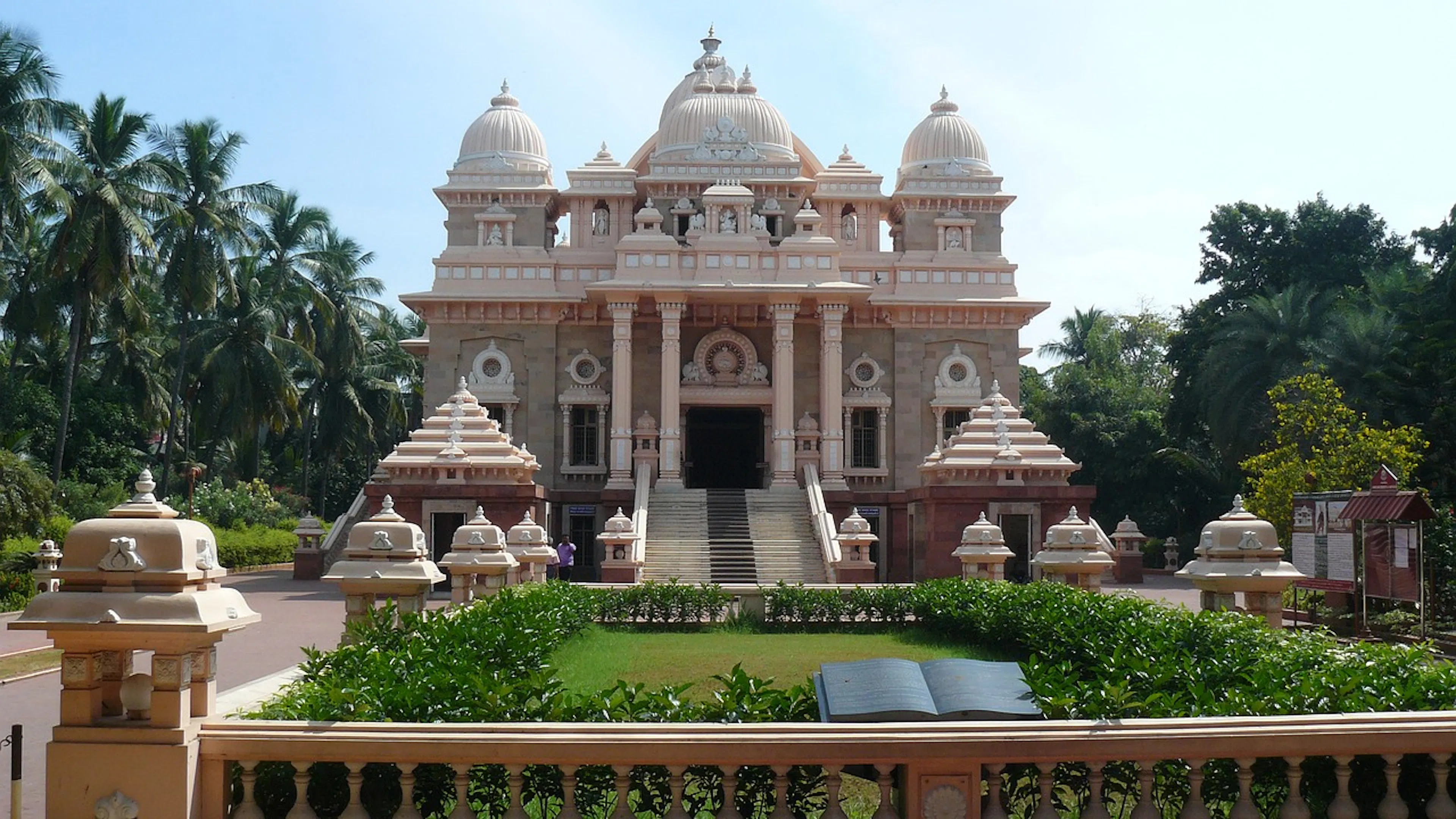 An elaborate Hindu temple built from a pink sandstone with pillars and domes and a lush garden stretching out in front