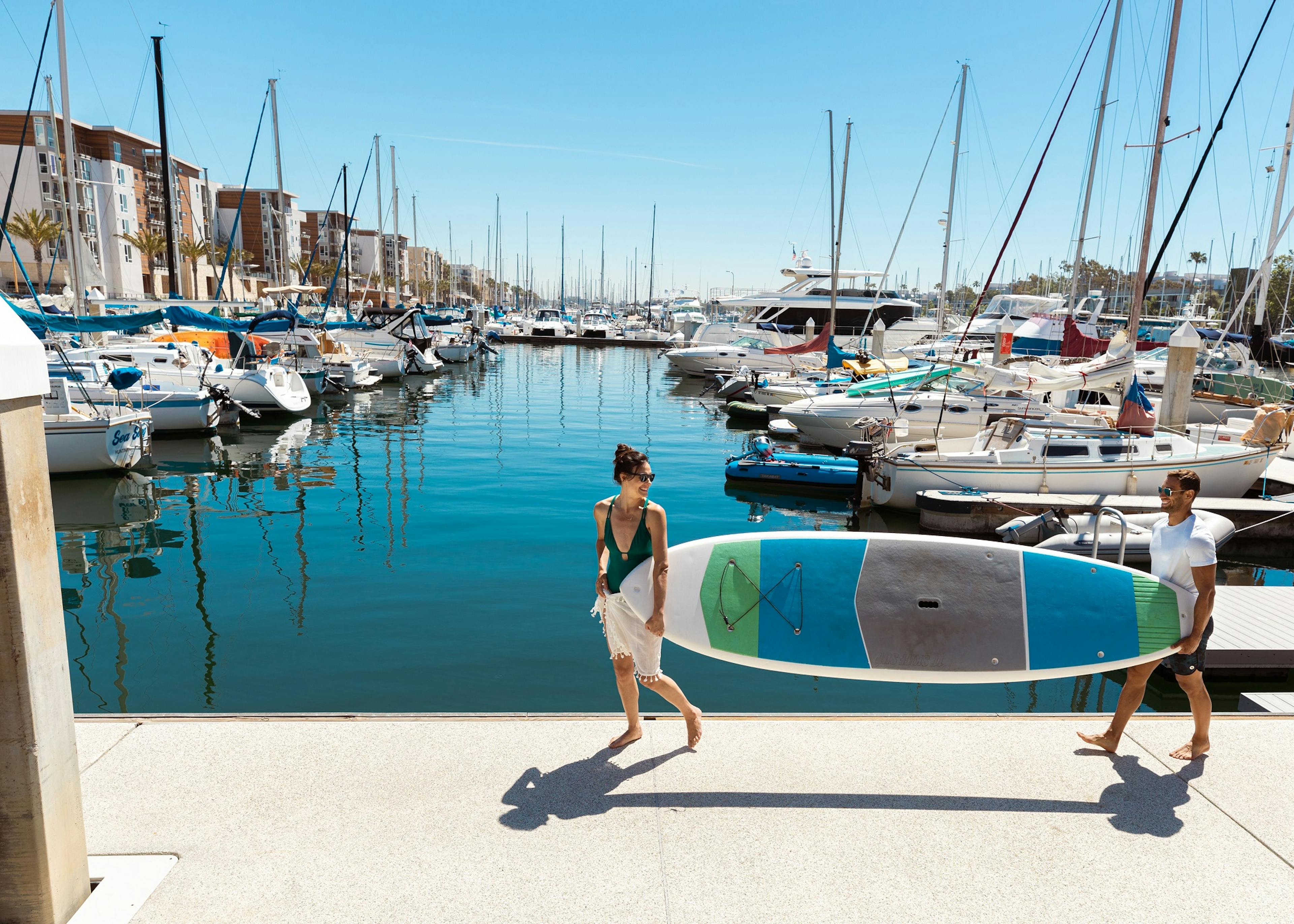 Two AMLI Marina Del Rey residents carrying a paddleboard with view of boats docked in front of the apartment community 
