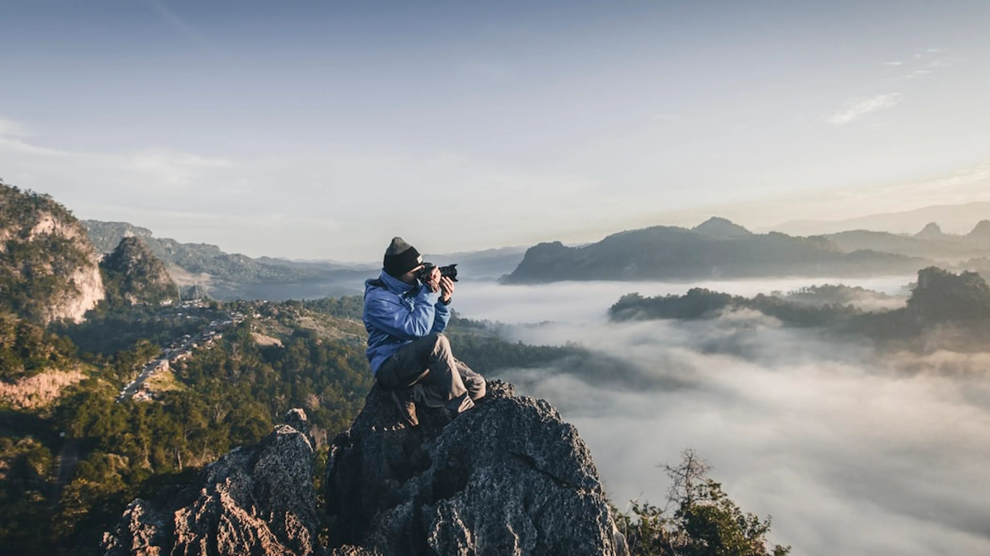 A man sitting on a mountain peak pointing a camera out into the distance