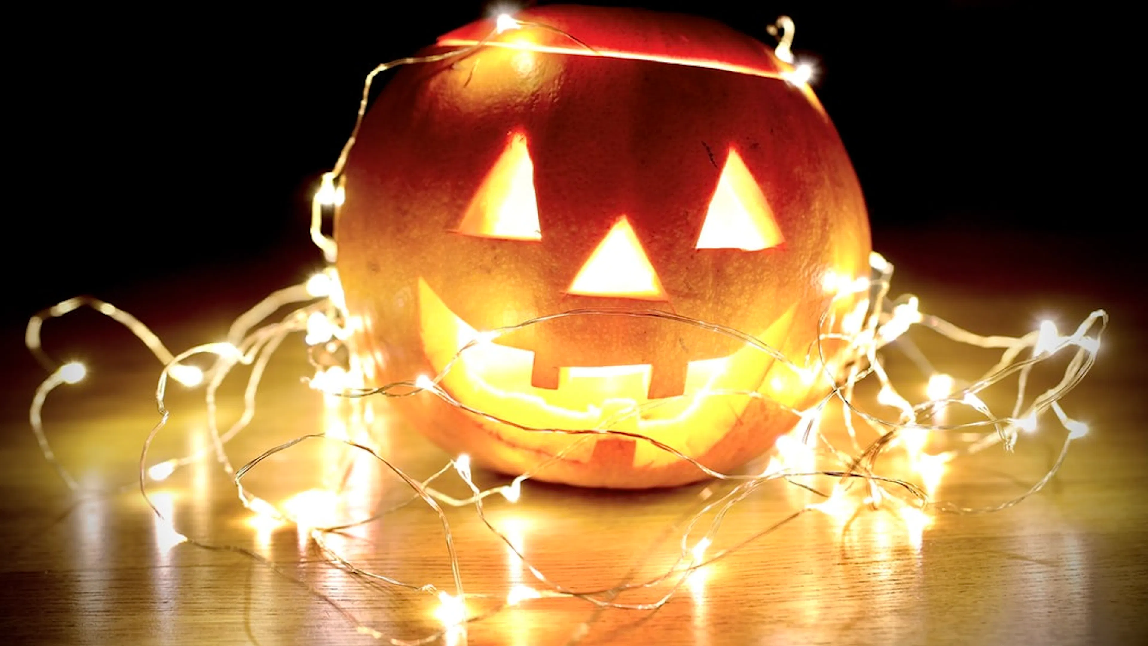 An orange Jack O Lantern with a smiley face and a candle shining from the inside surrounded by little string lights