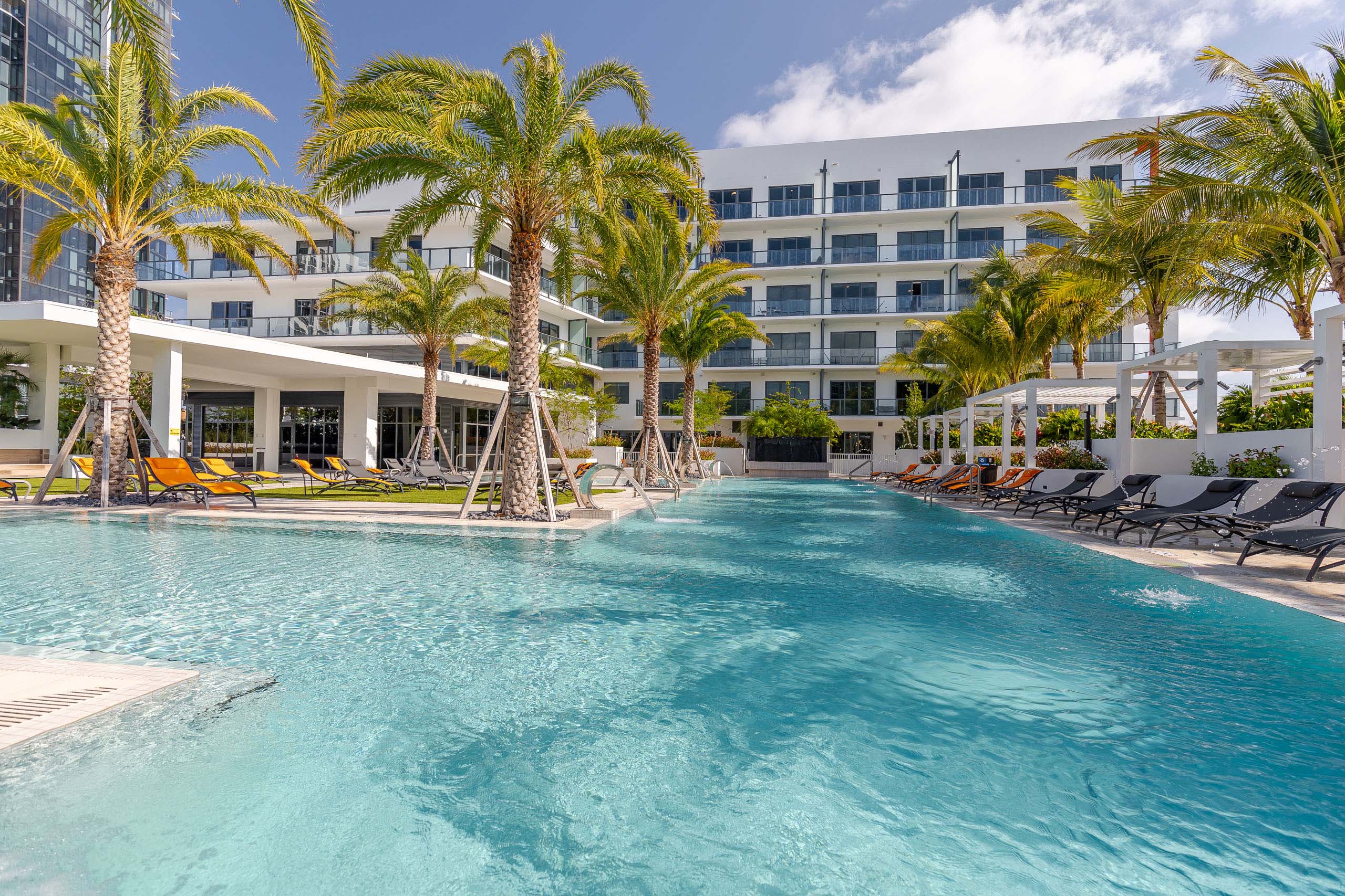 Pool at AMLI Midtown Miami apartments with clear blue water and lounge chairs and palm trees in front of of white building 