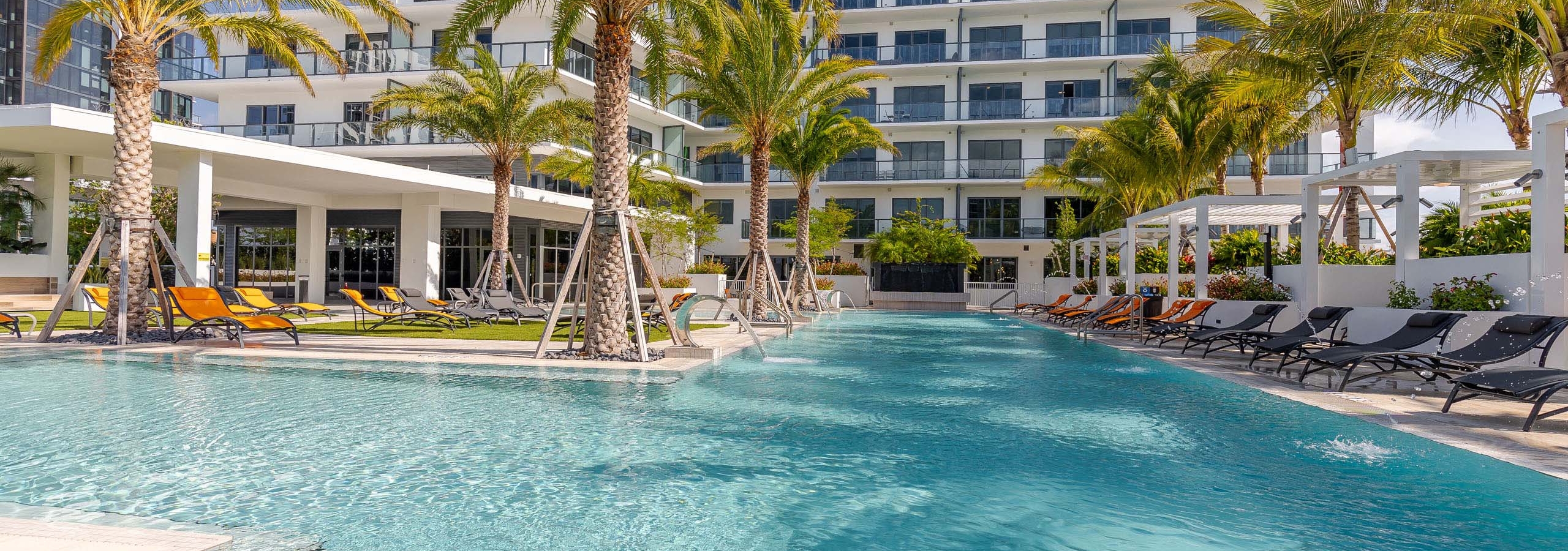 Pool at AMLI Midtown Miami apartments with clear blue water and lounge chairs and palm trees in front of of white building