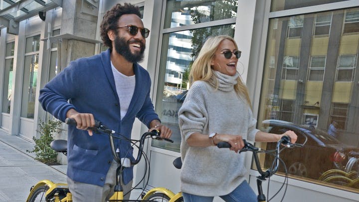 Two individuals wearing sunglasses and smiling riding bikes on a Denver sidewalk with concrete buildings in the background
