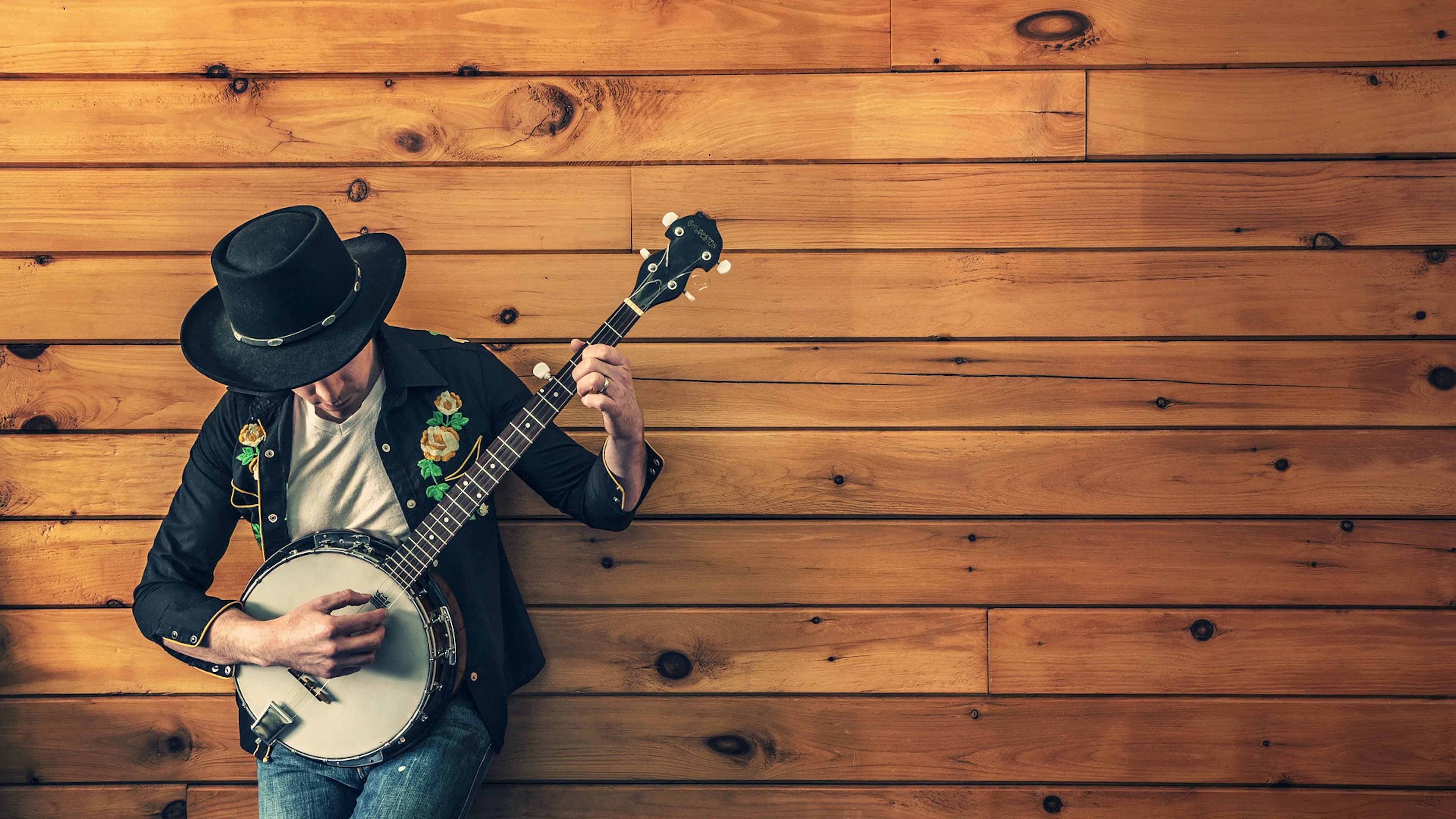 man in black hat playing banjo against pine wood wall