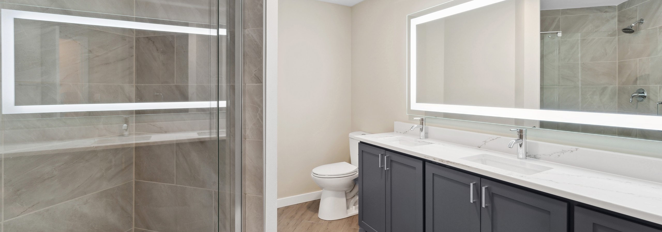 Bathroom at AMLI 535 with white quartz countertop on blue double vanity below lit mirror and glass shower with tile surround