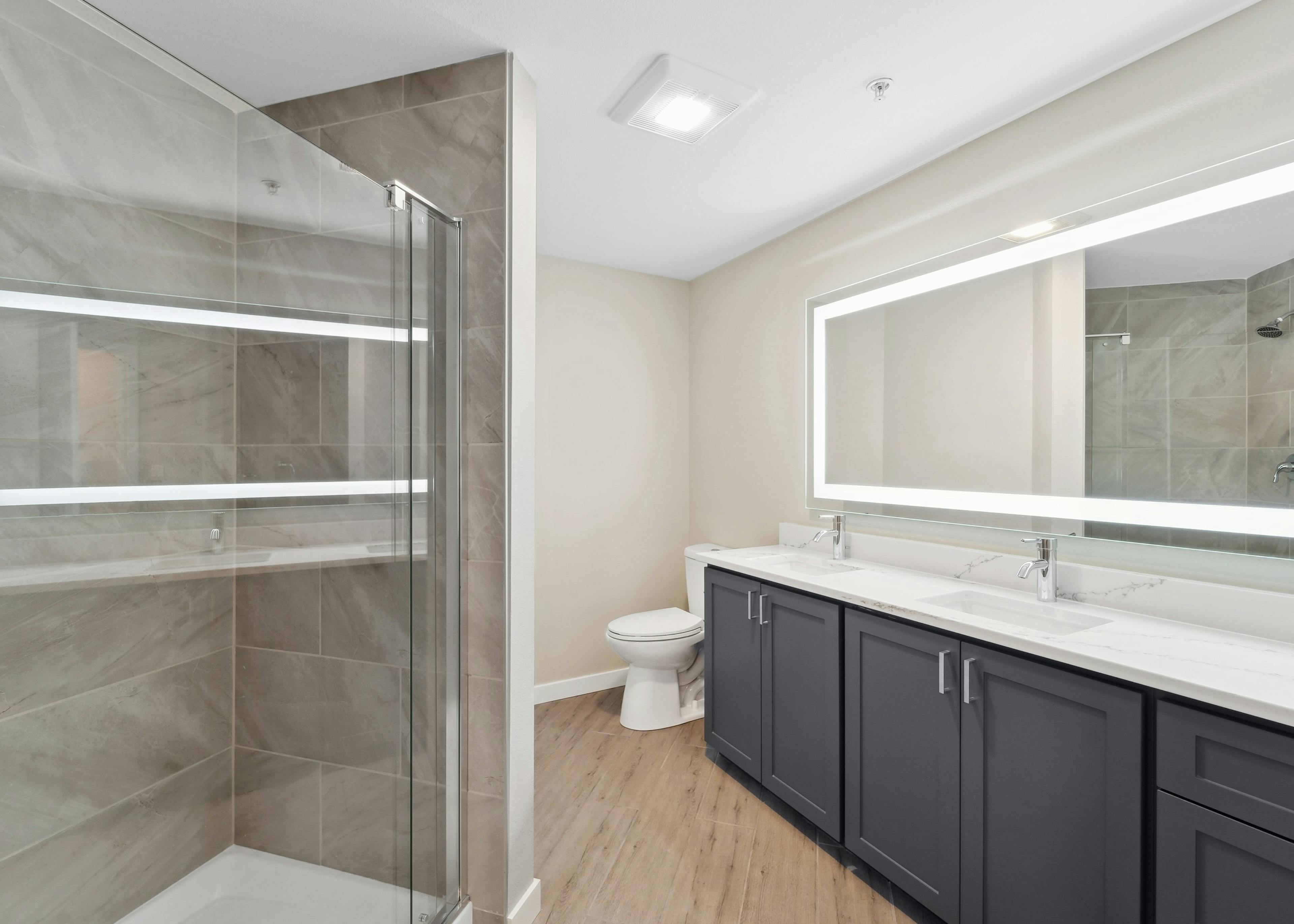 Bathroom at AMLI 535 with white quartz countertop on blue double vanity below lit mirror and glass shower with tile surround
