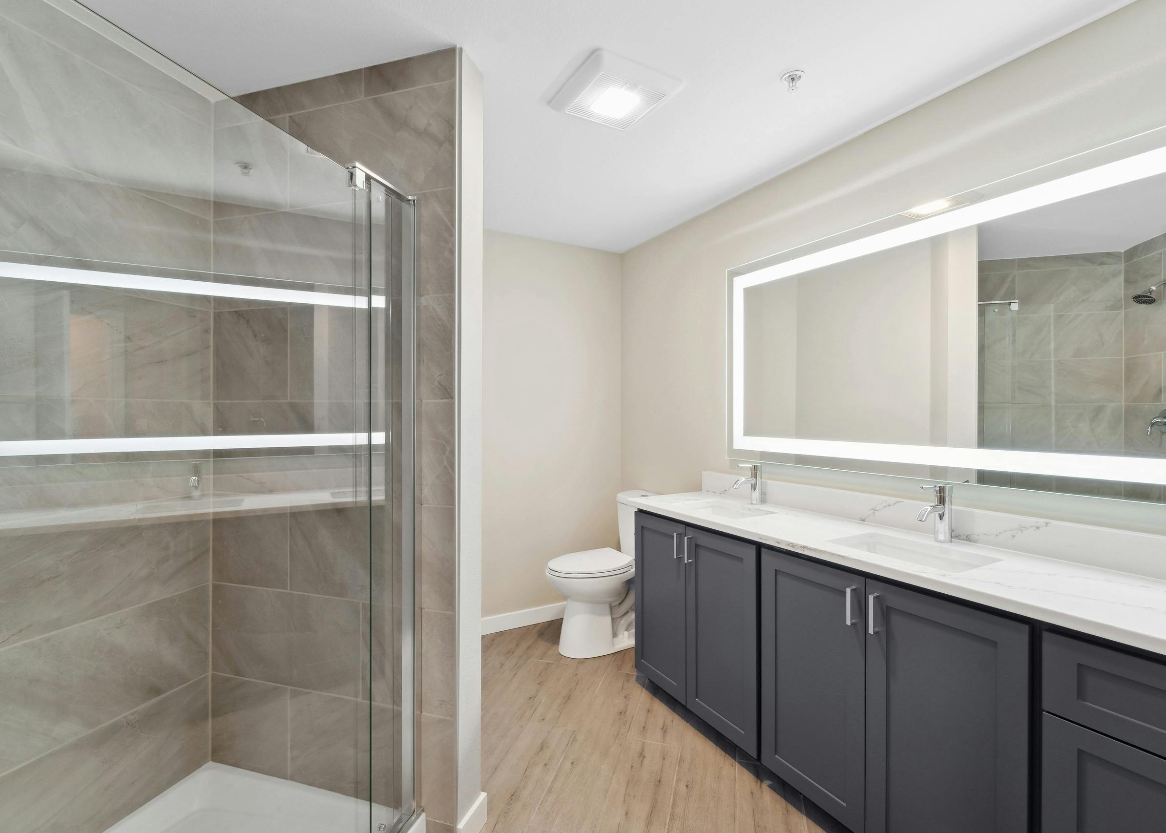 Bathroom at AMLI 535 with white quartz countertop on blue double vanity below lit mirror and glass shower with tile surround