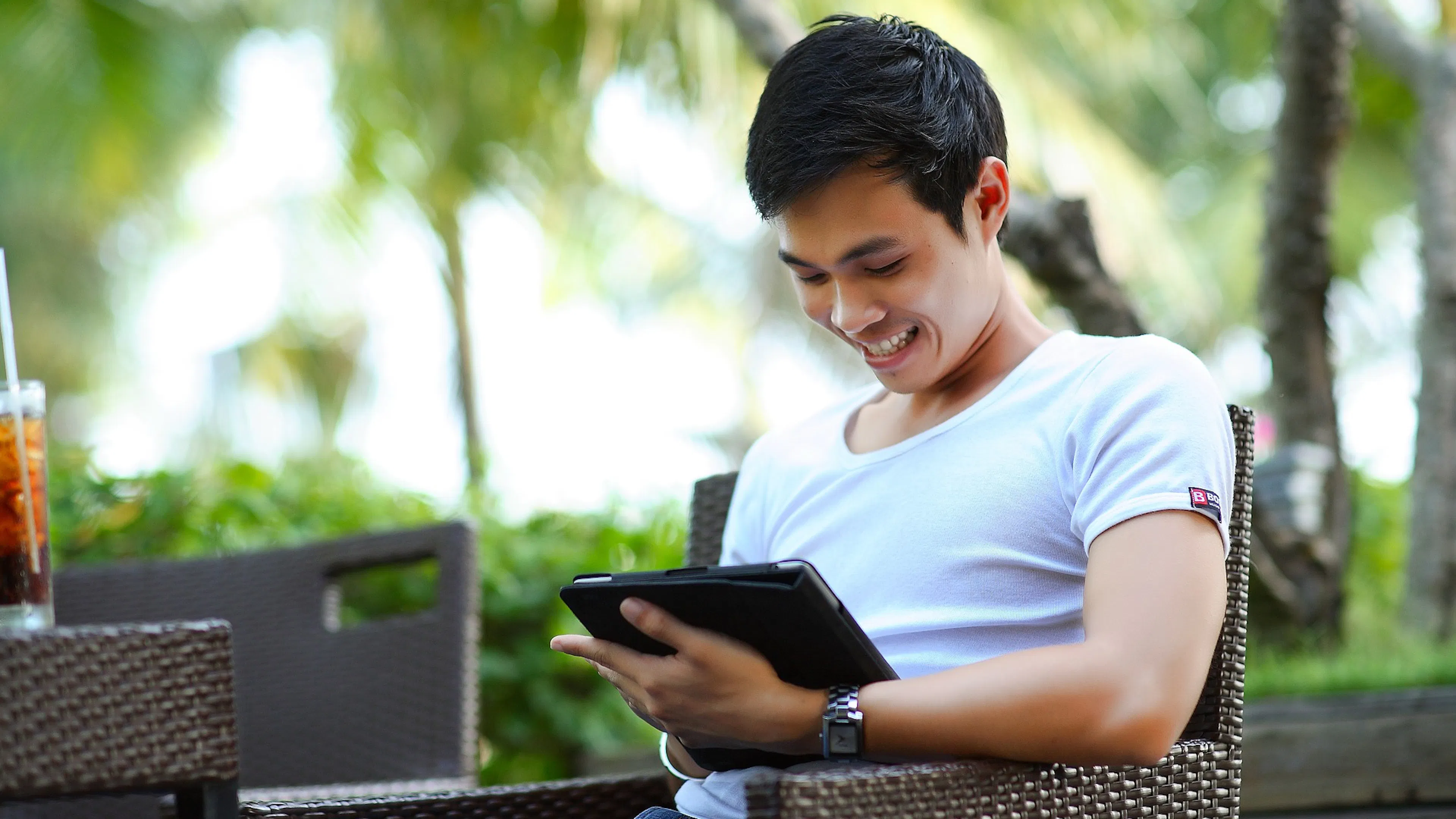 young man in white tshirt sitting in brown woven chair viewing tablet