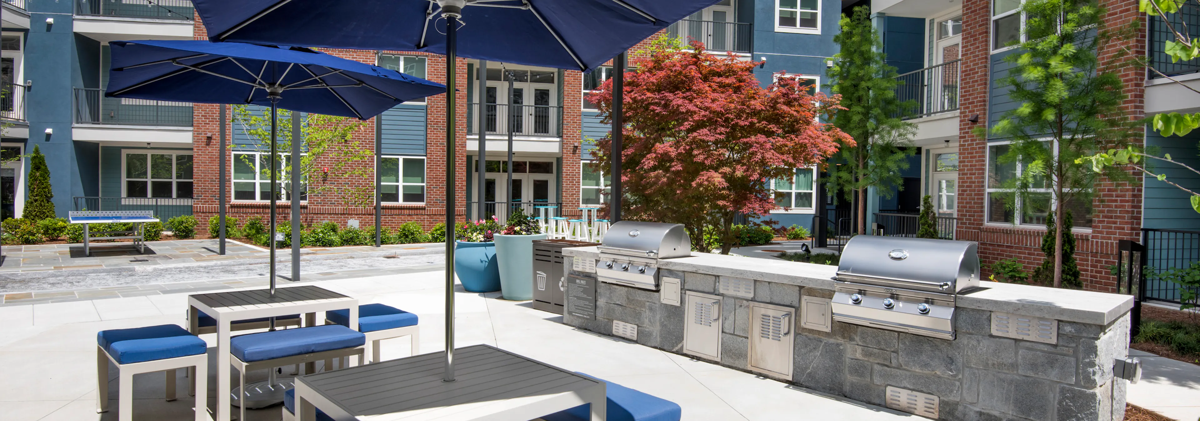 Daytime view of AMLI Decatur poolside grill area with two stainless grills and two tables with blue umbrellas and chairs