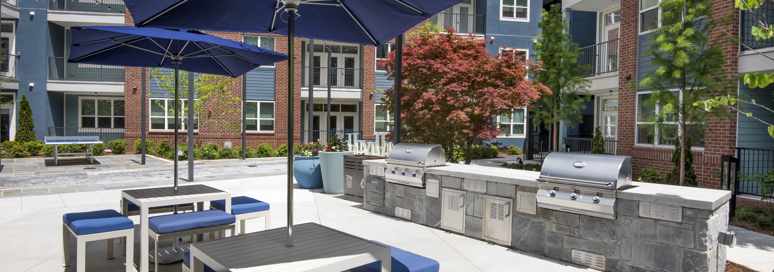 Daytime view of AMLI Decatur poolside grill area with two stainless grills and two tables with blue umbrellas and chairs