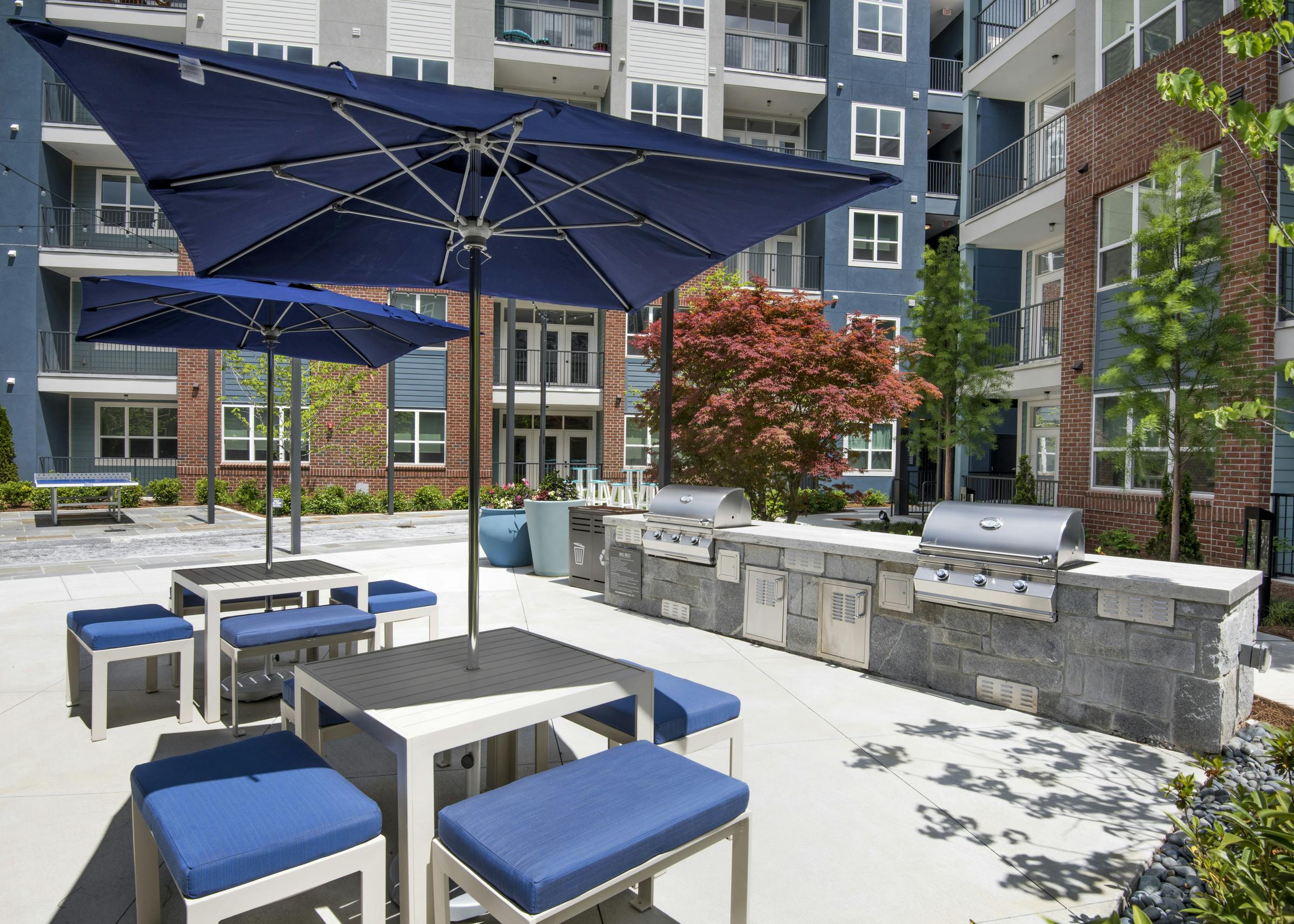 Daytime view of AMLI Decatur poolside grill area with two stainless grills and two tables with blue umbrellas and chairs