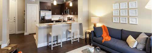 Interior of living room and kitchen at AMLI West Plano with a dark grey couch and tan barstools at a granite counter