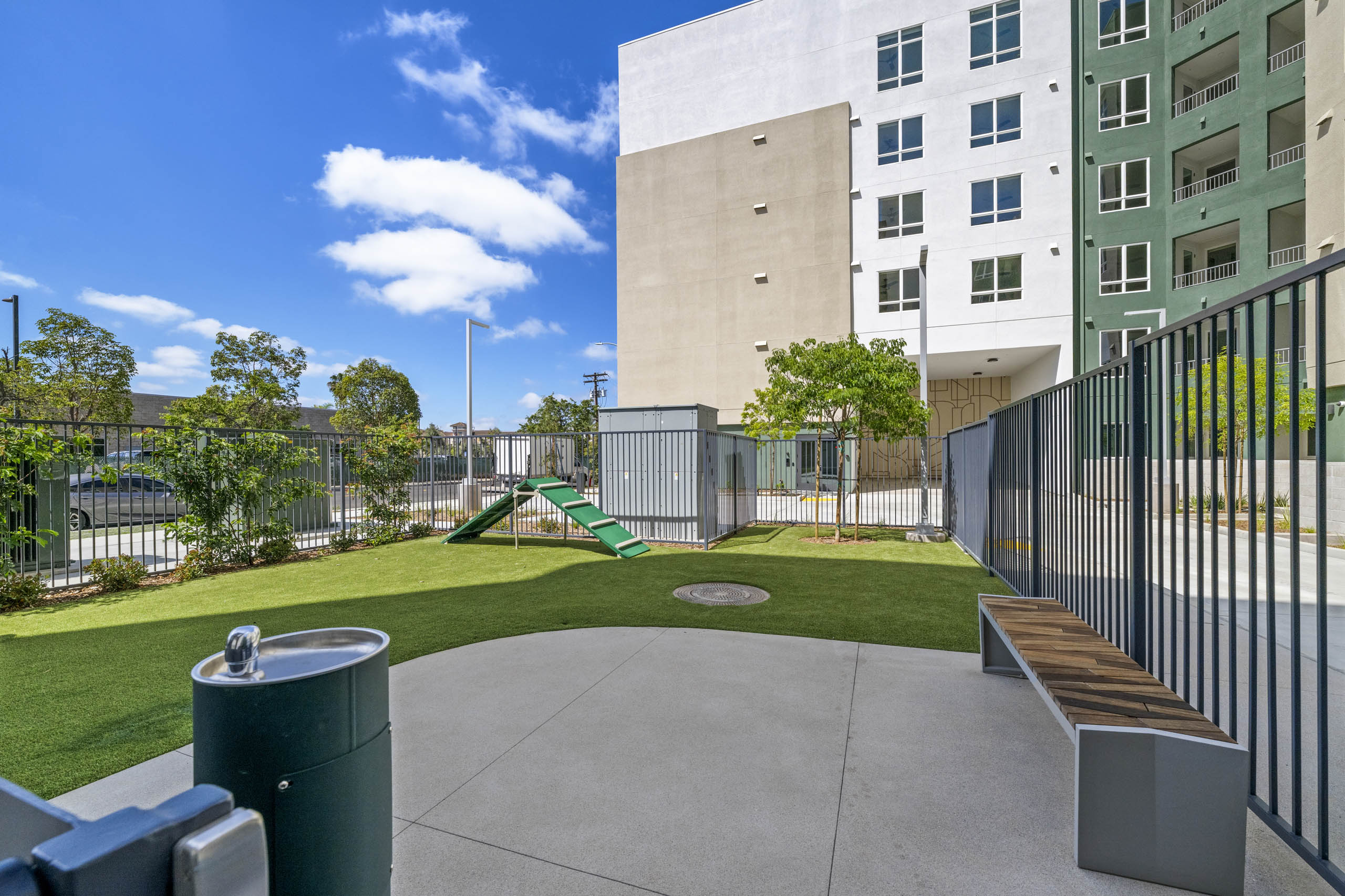 Fenced dog park with artificial turf, agility ramp, bench seating, water fountain, landscaped greenery.