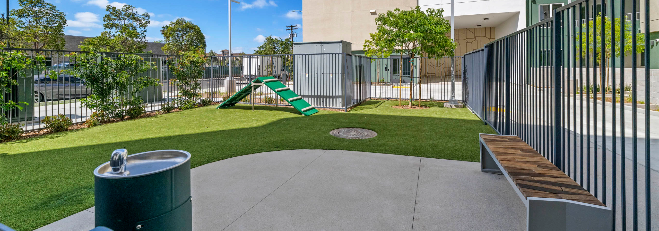 Fenced dog park with artificial turf, agility ramp, bench seating, water fountain, landscaped greenery.