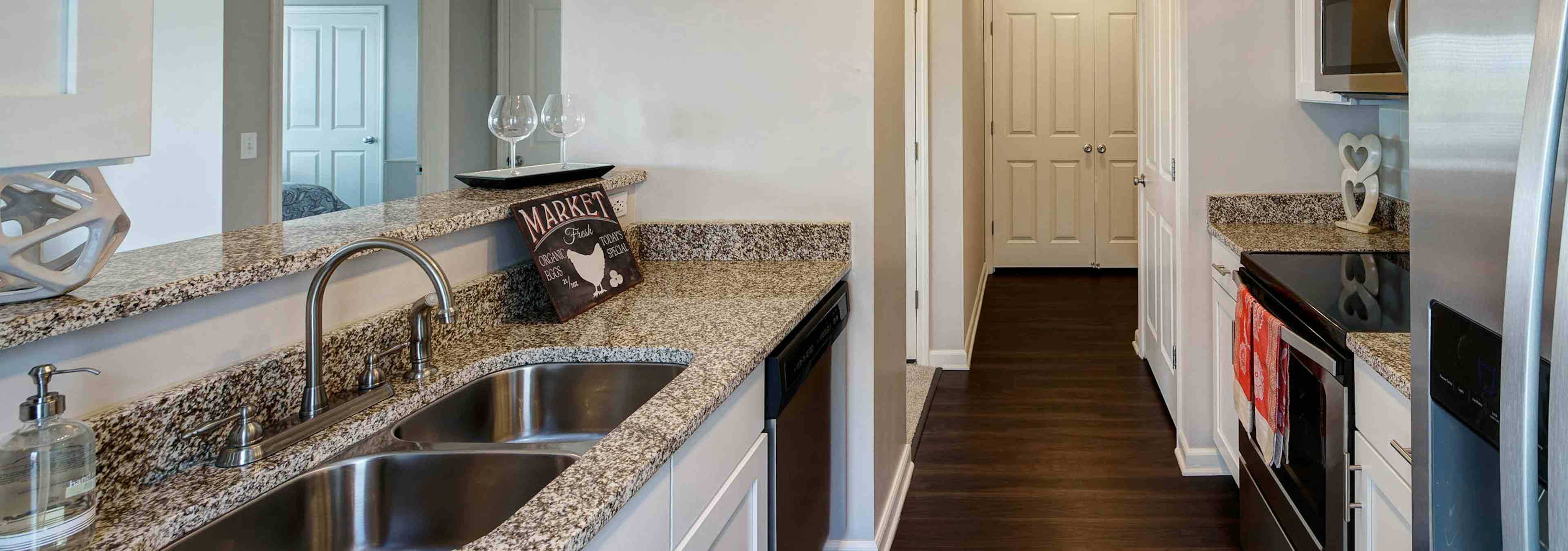 AMLI at Seven Bridges kitchen with white cabinets and granite countertops with dark wood floors leading to a hallway 