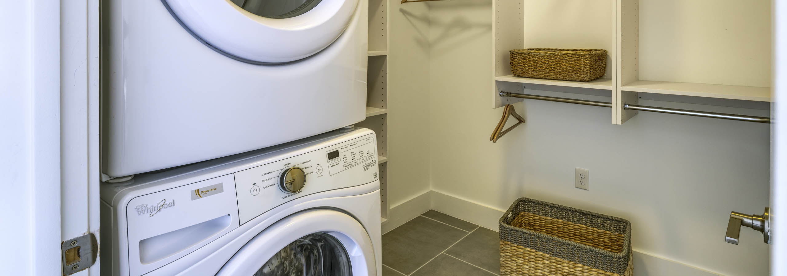 Laundry room with stacked white washer and dryer and white shelves with wood  hangers and wicker baskets at AMLI Midtown 29
