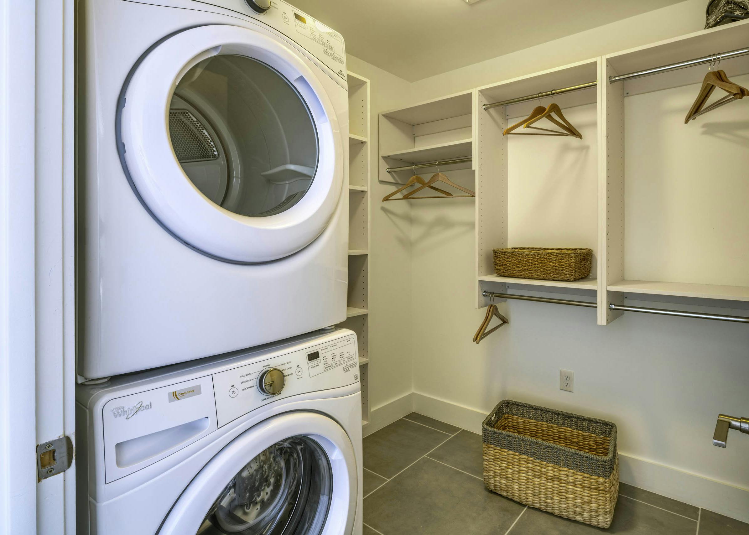Laundry room with stacked white washer and dryer and white shelves with wood hangers and wicker baskets at AMLI Midtown 29