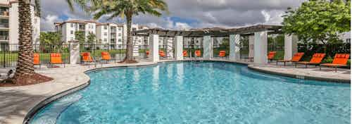 Daytime view of AMLI Doral swimming pool surrounded by bright orange lounge chairs, palm trees and freestanding pergola