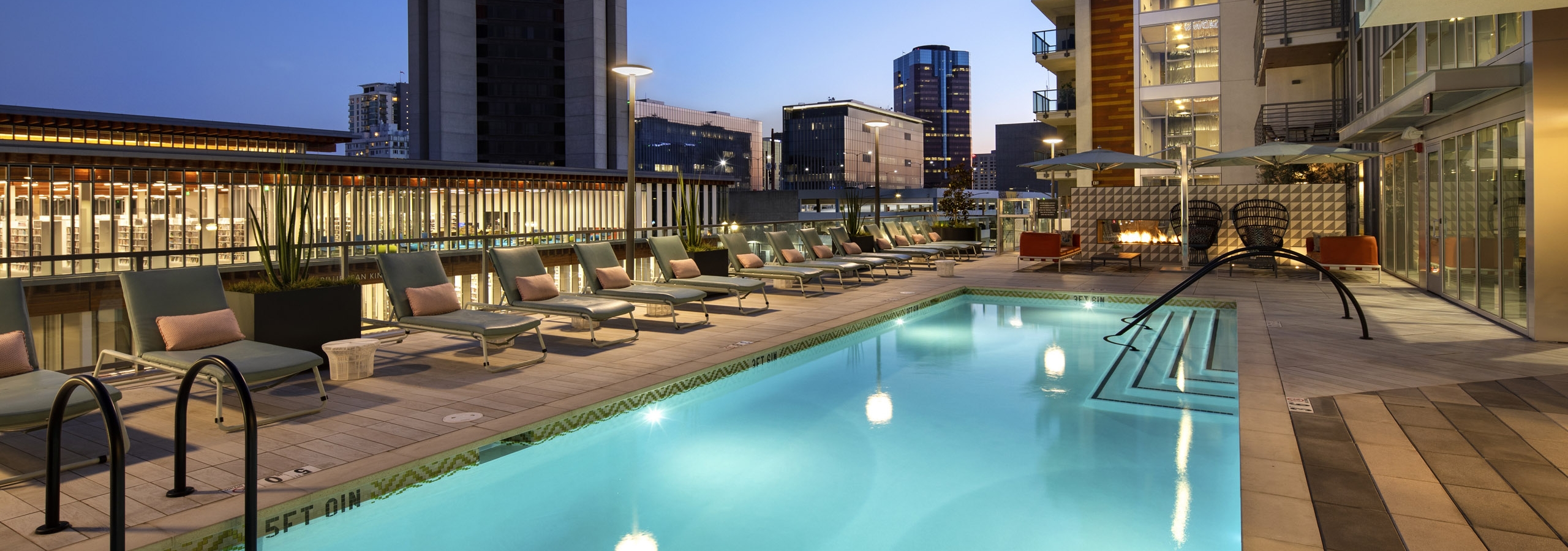 Nighttime view of AMLI Park Broadway swimming pool area with lounge seating overlooking library and Downtown Long Beach