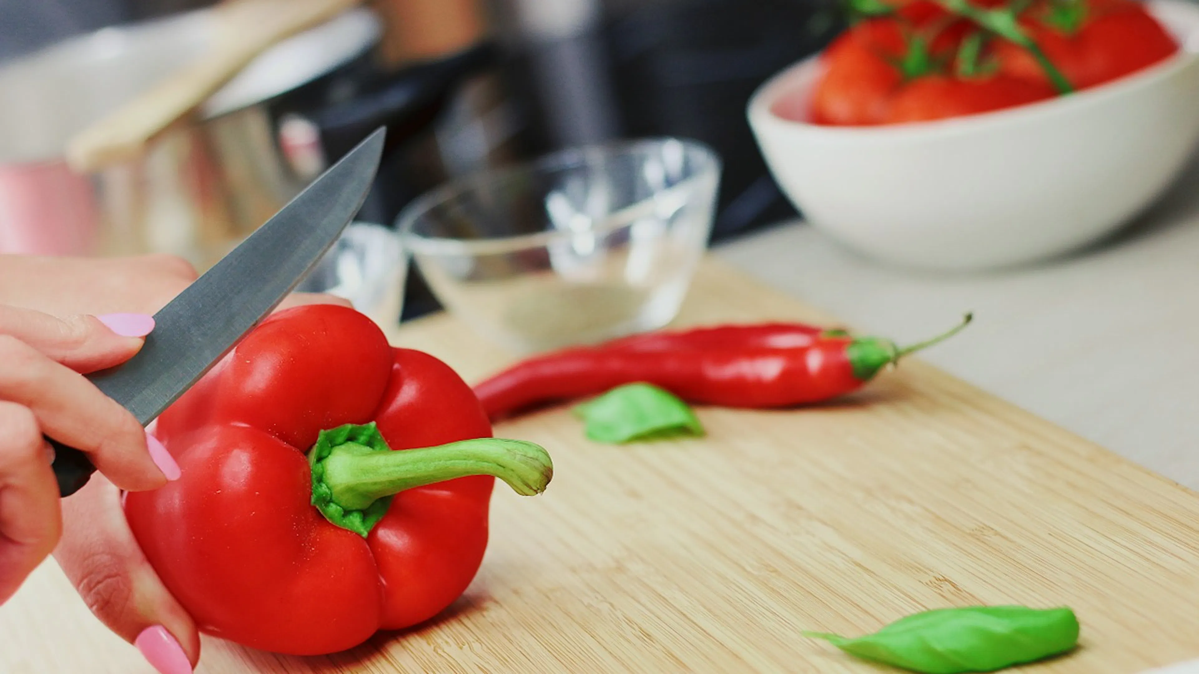 Woman with pink fingernails slicing a red pepper on a bamboo wooden cutting board with other peppers in the background