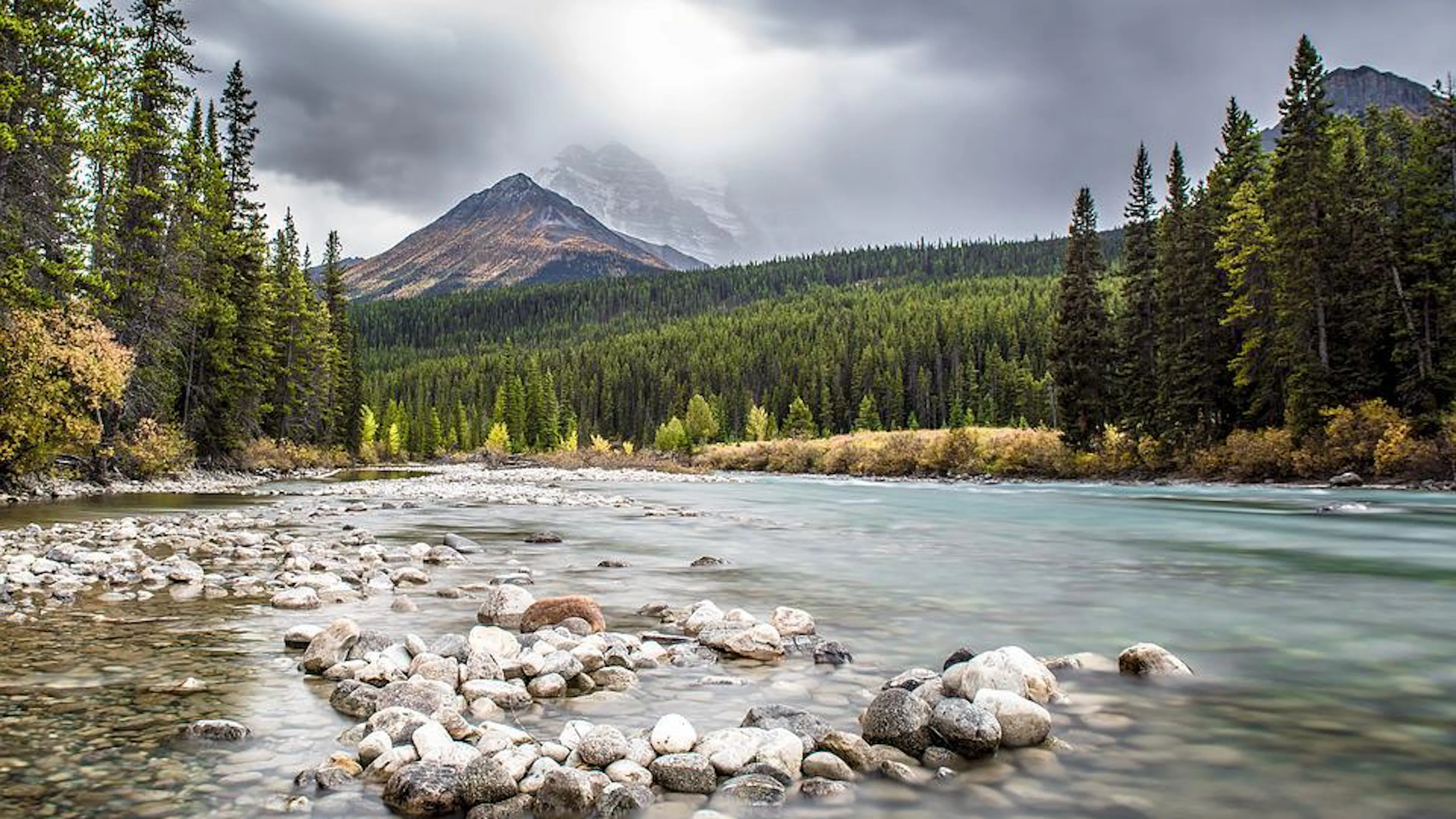 A clear river winding below a tall forested mountain on a cloudy day