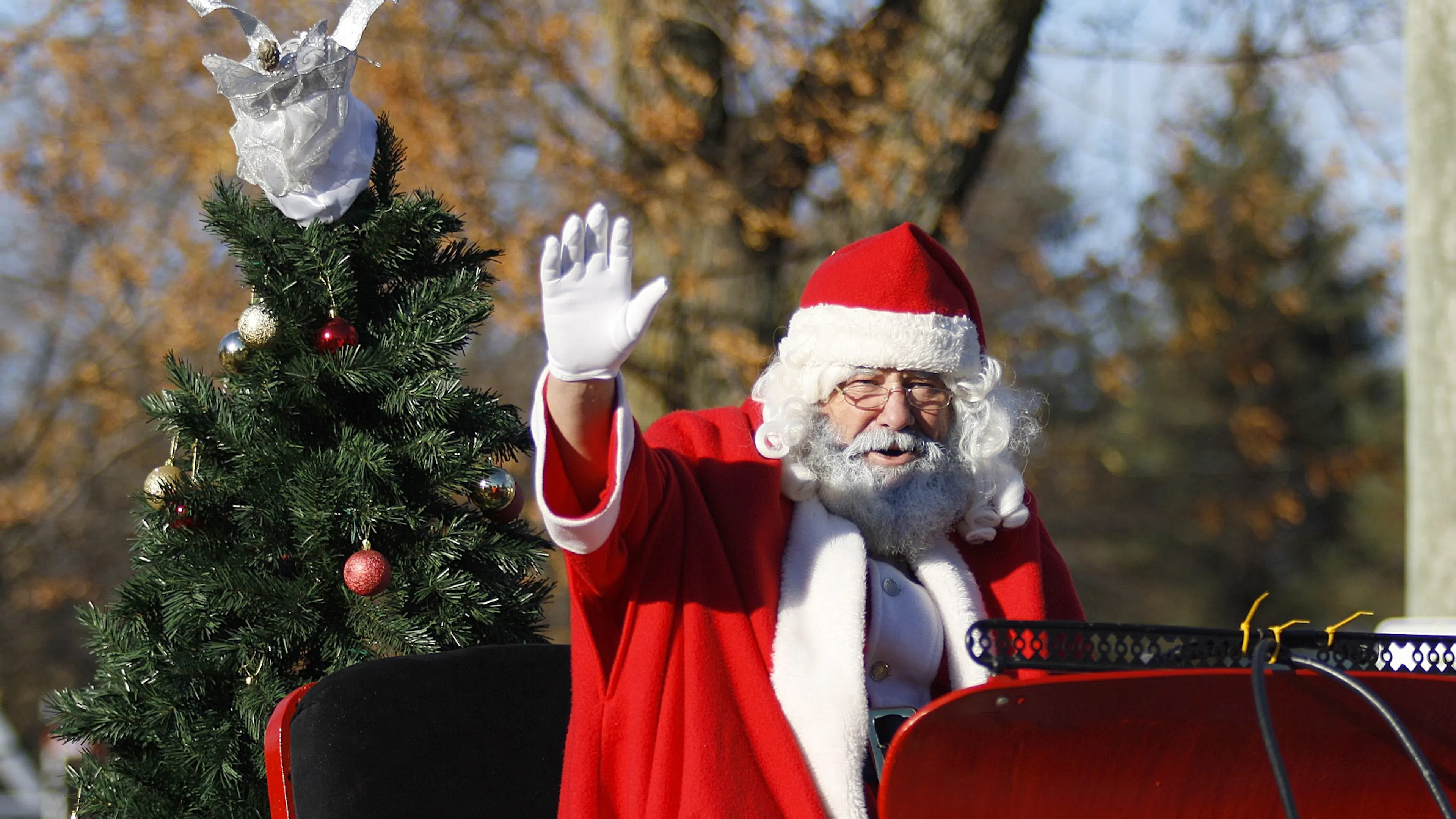 Santa Claus in a red sleigh waving to the unseen crowd with a small Christmas tree on the back of the sleigh
