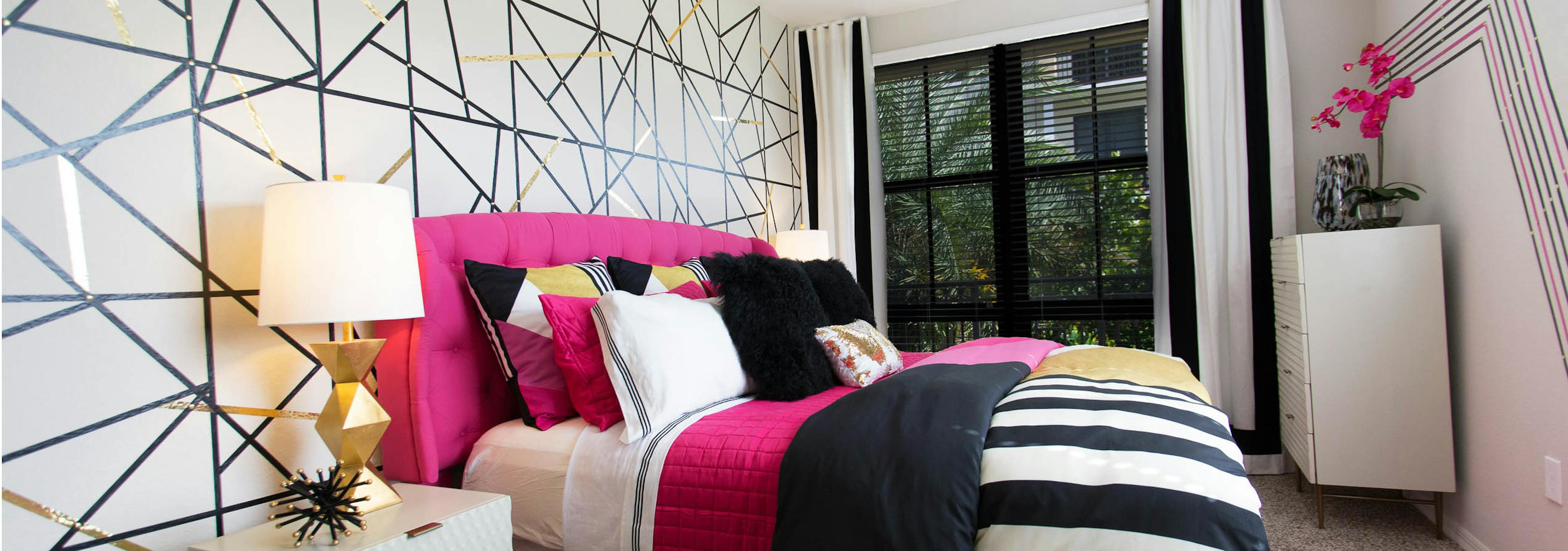 AMLI Dadeland master bedroom with geometric accent wall, modern table lamp, magenta and black bedding and a window view