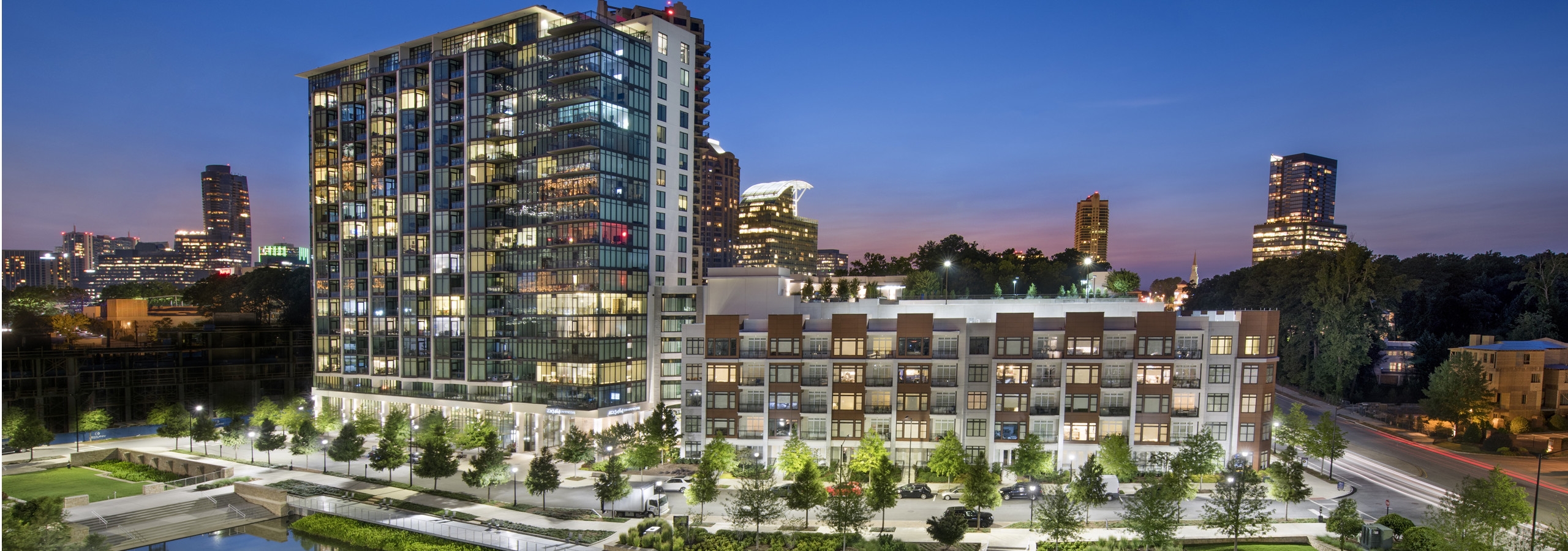 Exterior of the AMLI 3464 building at night which is filled with lit windows and overlooks bright trees lining the pavement