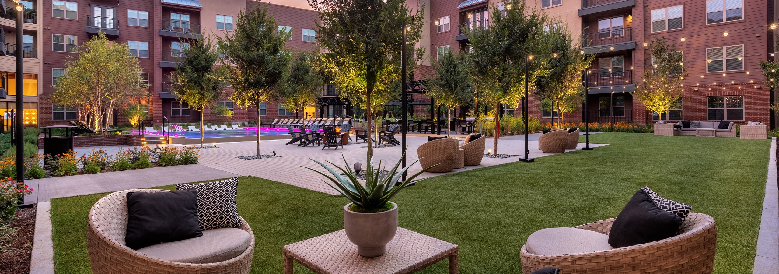 Dusk view of AMLI Grapevine outdoor living room with wicker chairs and tables on grass in courtyard with pool in background