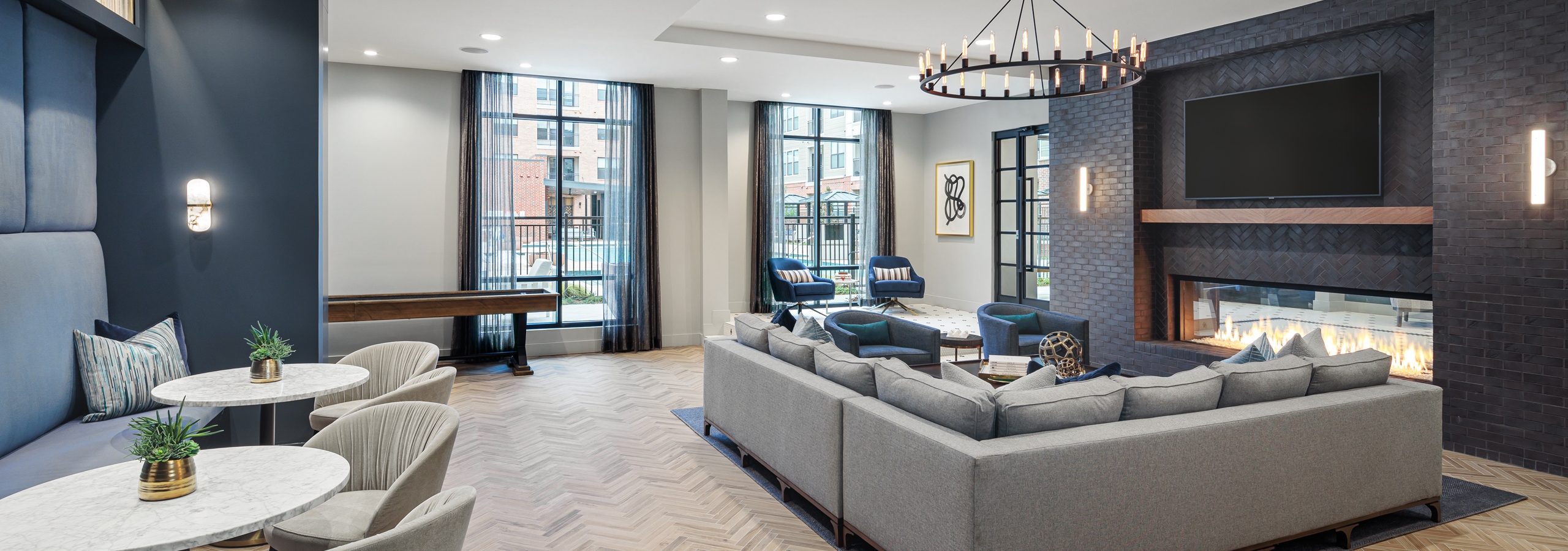 AMLI Addison resident parlor with long grey couch in front of a fireplace with tables, chairs and large windows in background