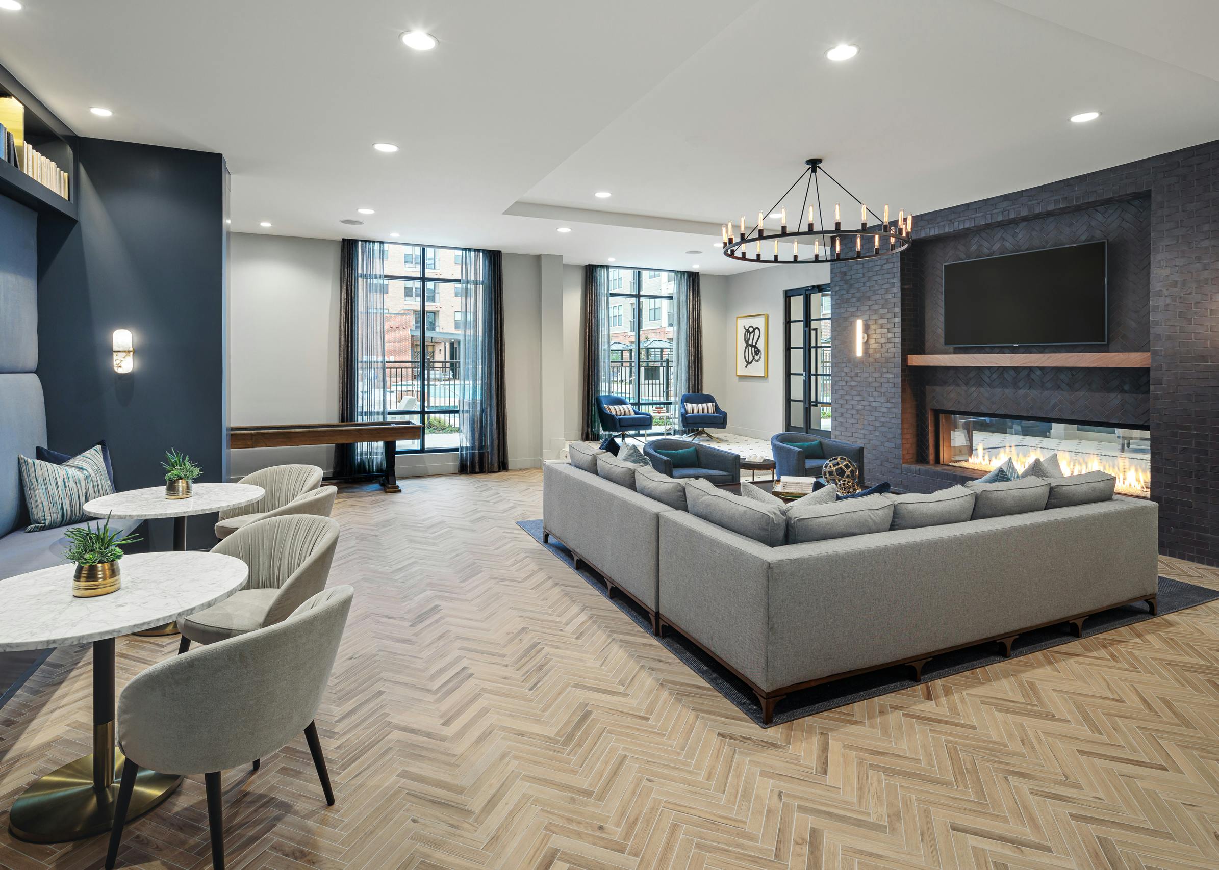 AMLI Addison resident parlor with long grey couch in front of a fireplace with tables, chairs and large windows in background