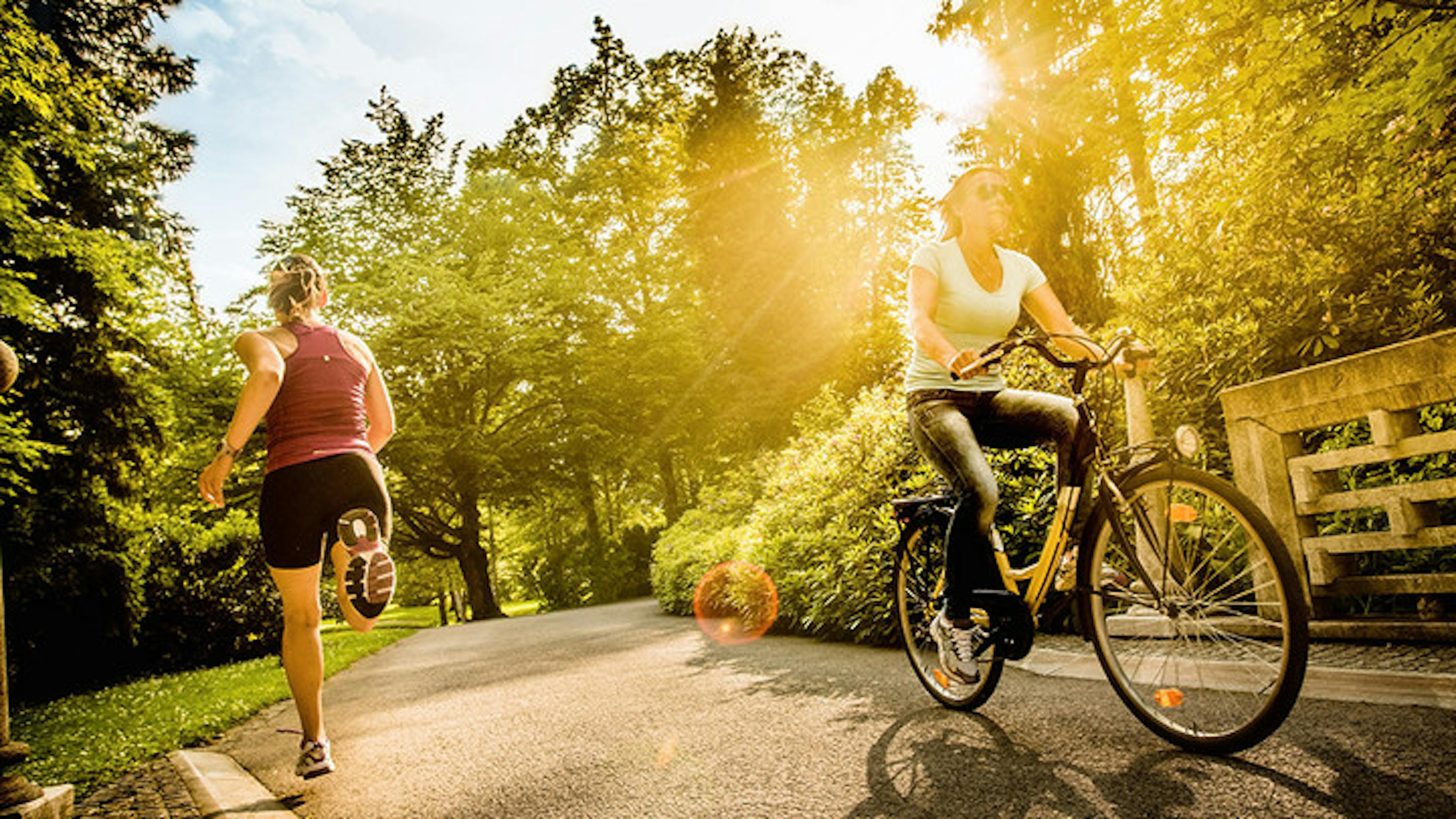 Runner and biker passing one another on a trail with green trees and bushes with rays of sunlight streaming through foliage