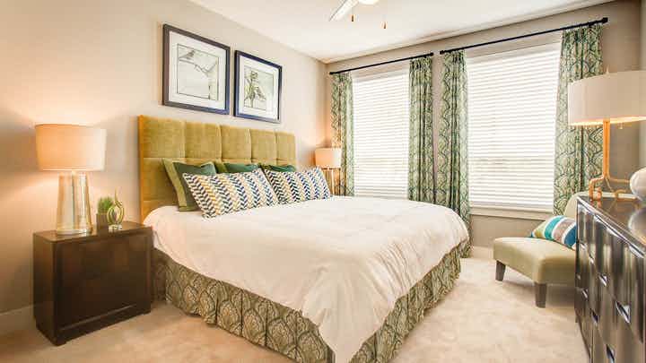 A bedroom at AMLI Riverfront Park apartments with lamps and artwork and a window with green curtains and a balcony view
