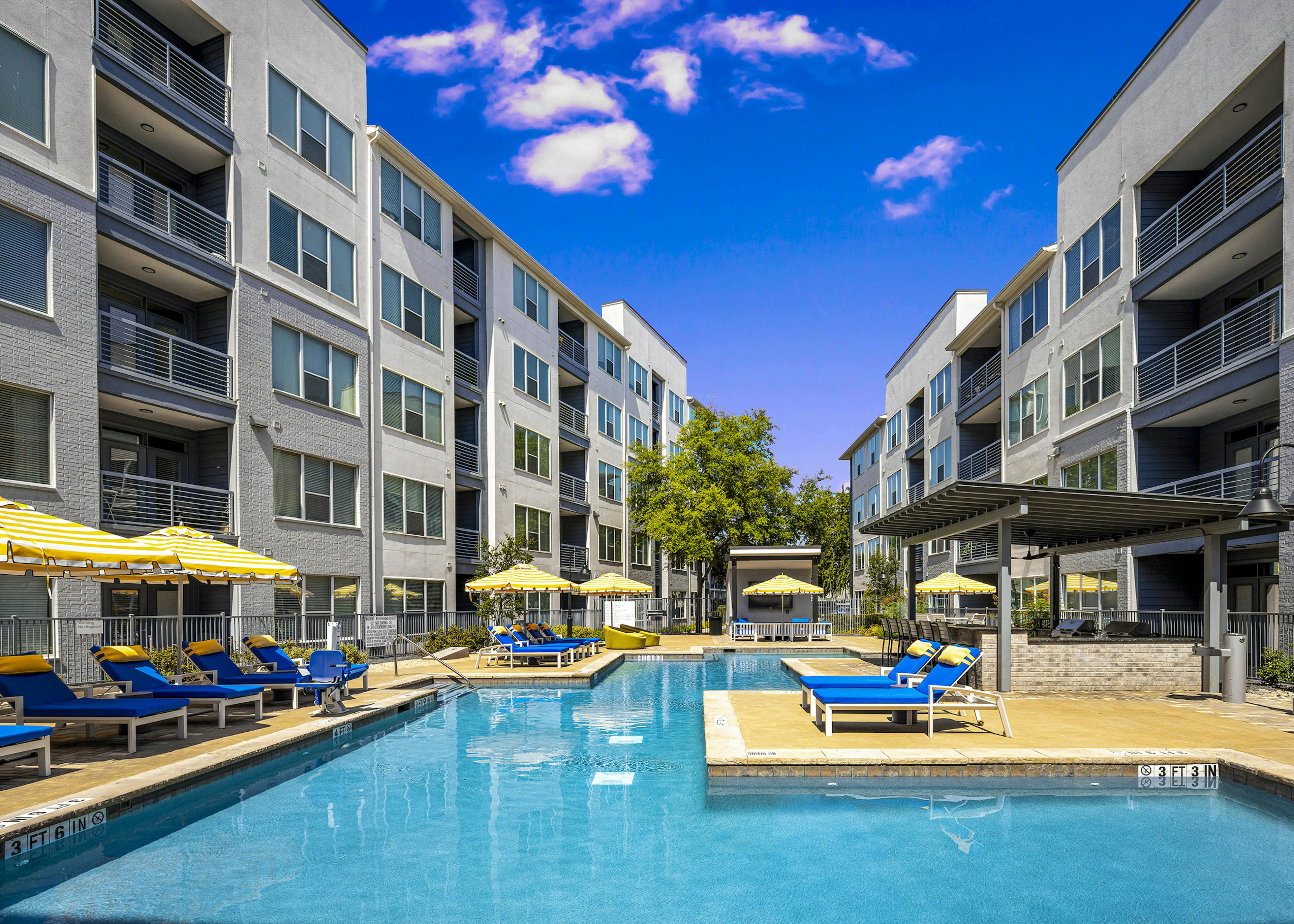 Daytime view of AMLI South Shore pool with surrounding blue lounge chairs and yellow and white umbrellas between 2 buildings