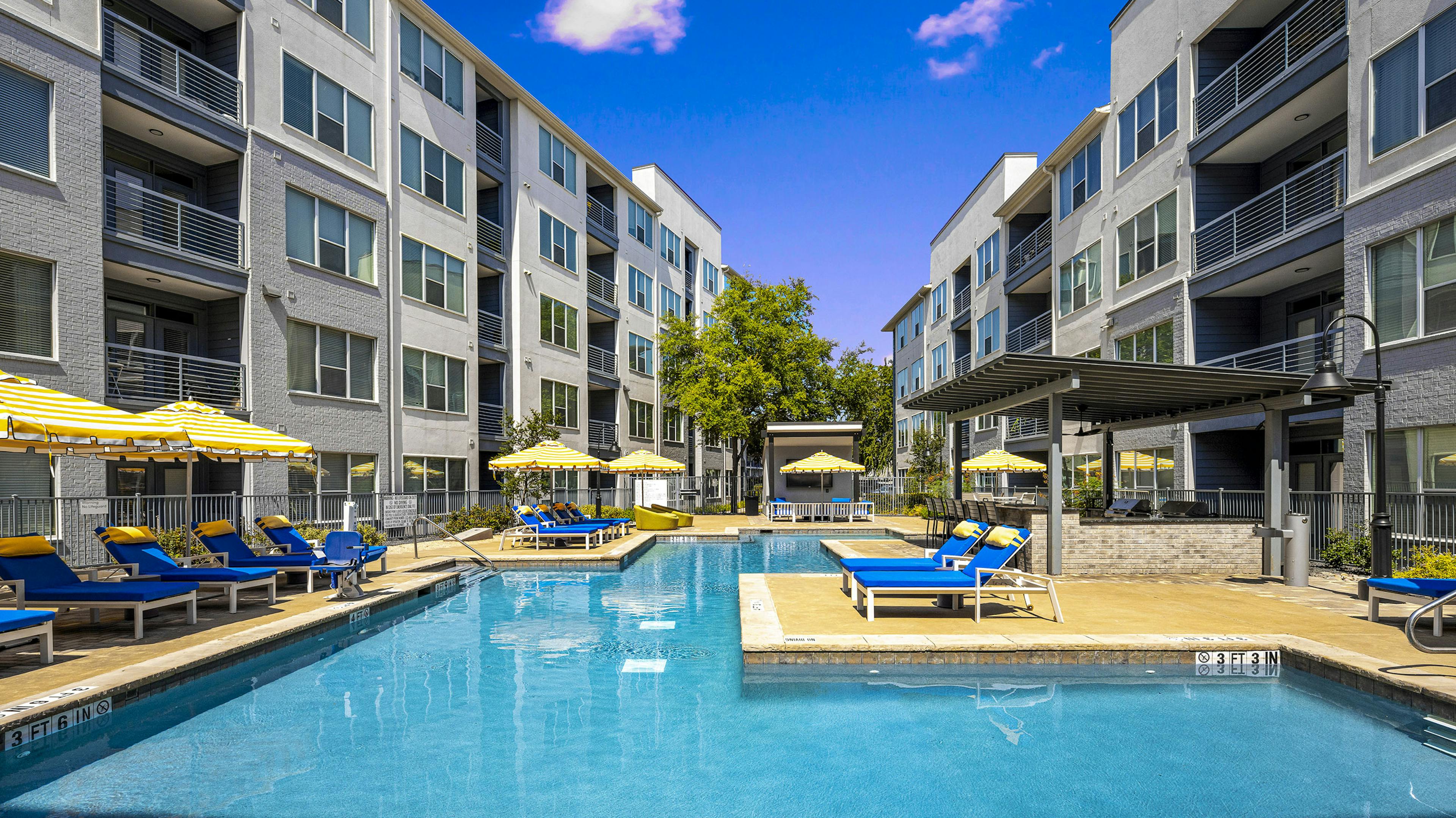 Daytime view of AMLI South Shore pool with surrounding blue lounge chairs and yellow and white umbrellas between 2 buildings