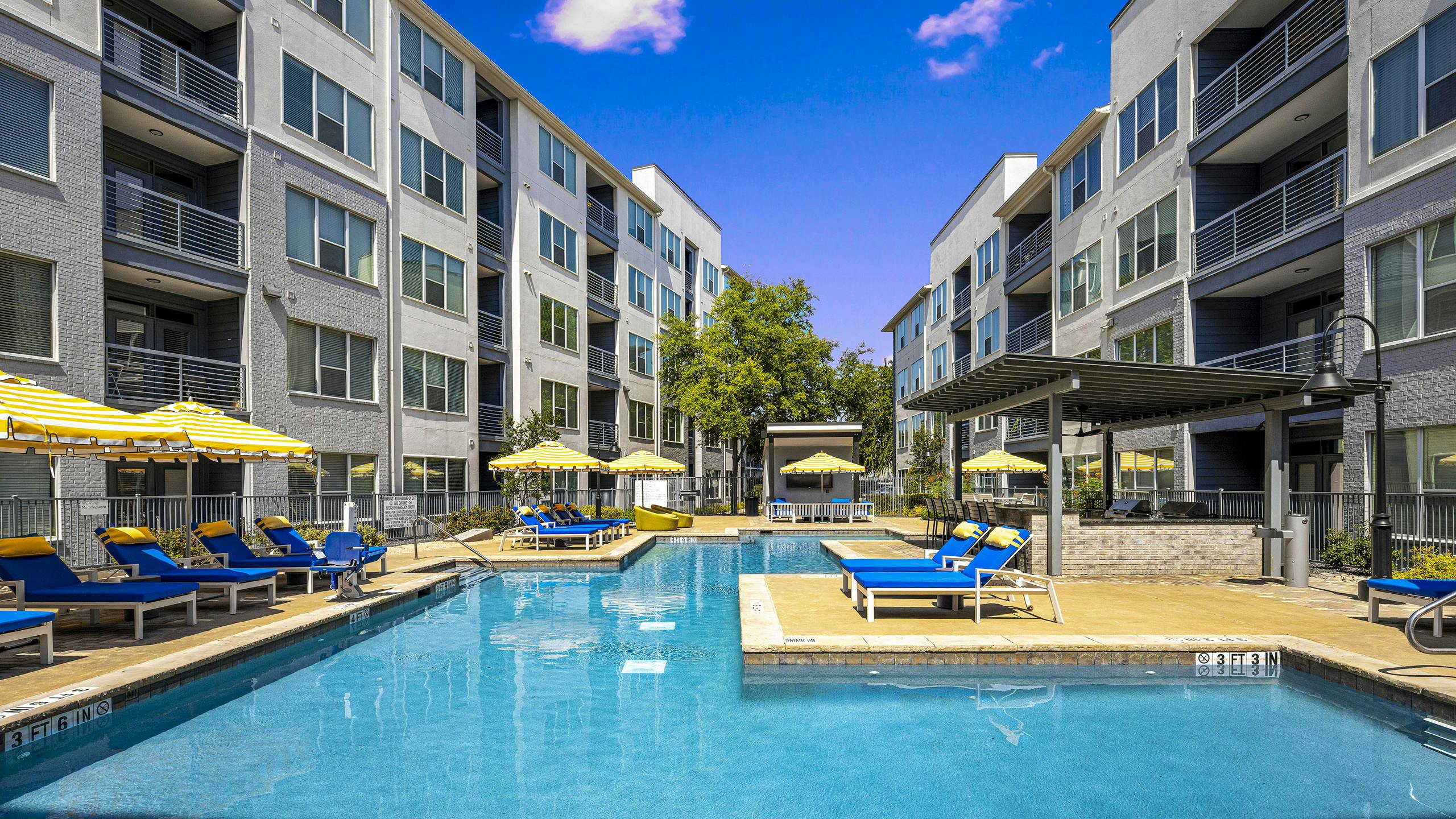 Daytime view of AMLI South Shore pool with surrounding blue lounge chairs and yellow and white umbrellas between 2 buildings