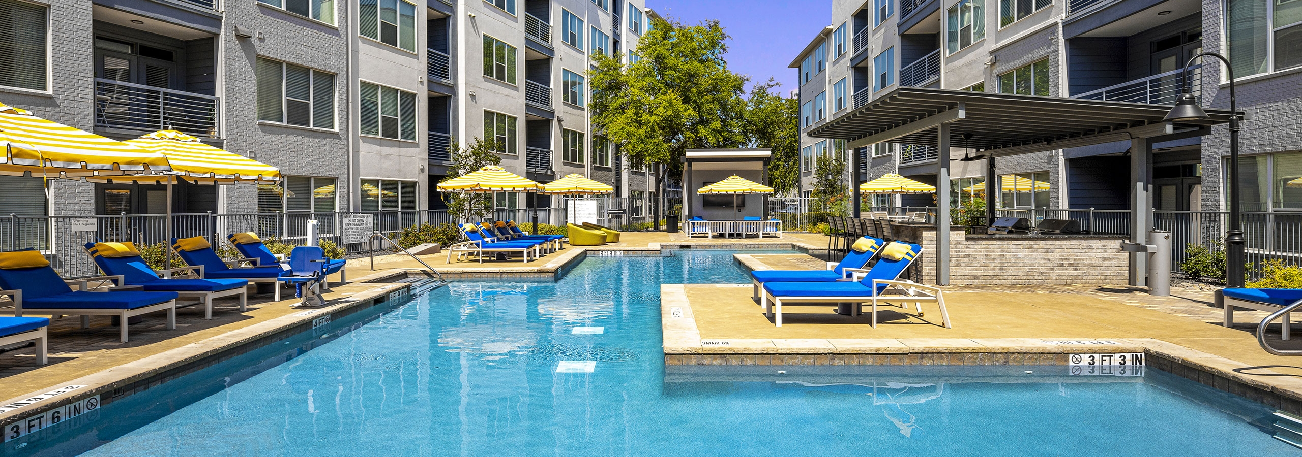 Daytime view of AMLI South Shore pool with surrounding blue lounge chairs and yellow and white umbrellas between 2 buildings