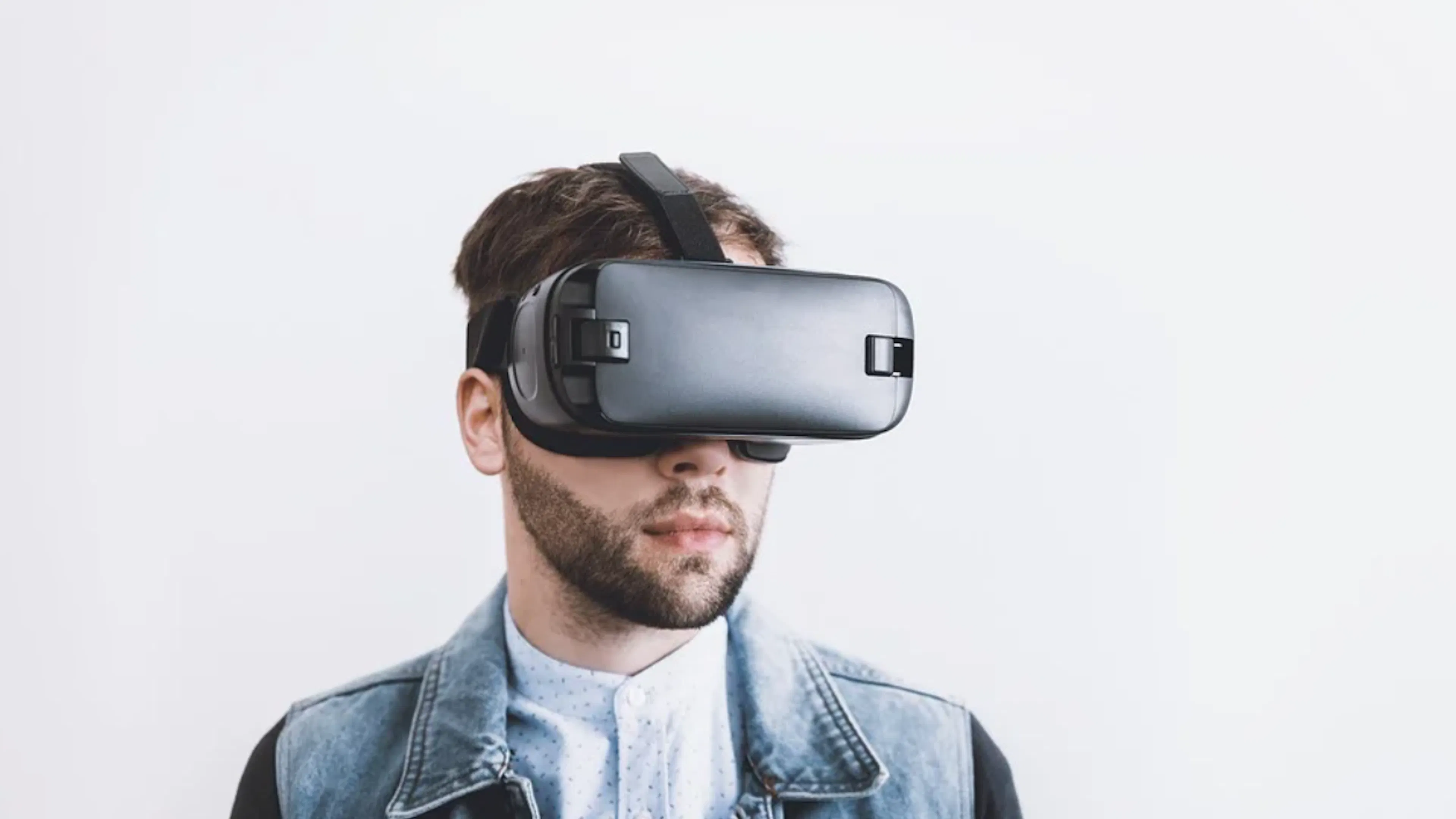 A man wearing a denim jacket and a virtual reality headset on against a bright white background