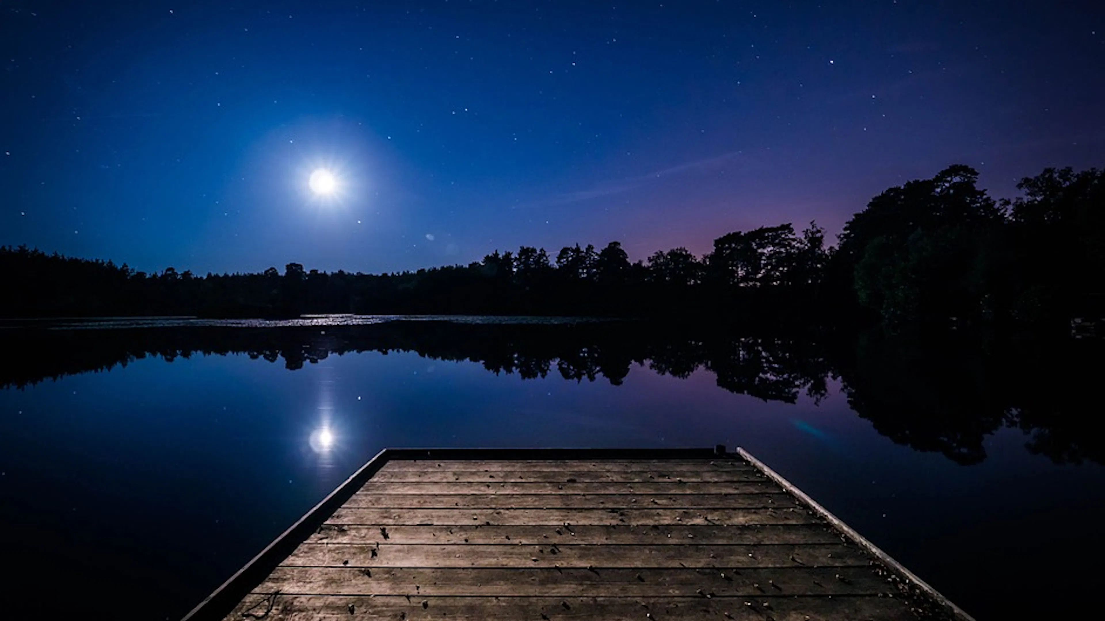 A jetty sticking out into a lake under a night sky with the moon shining bright