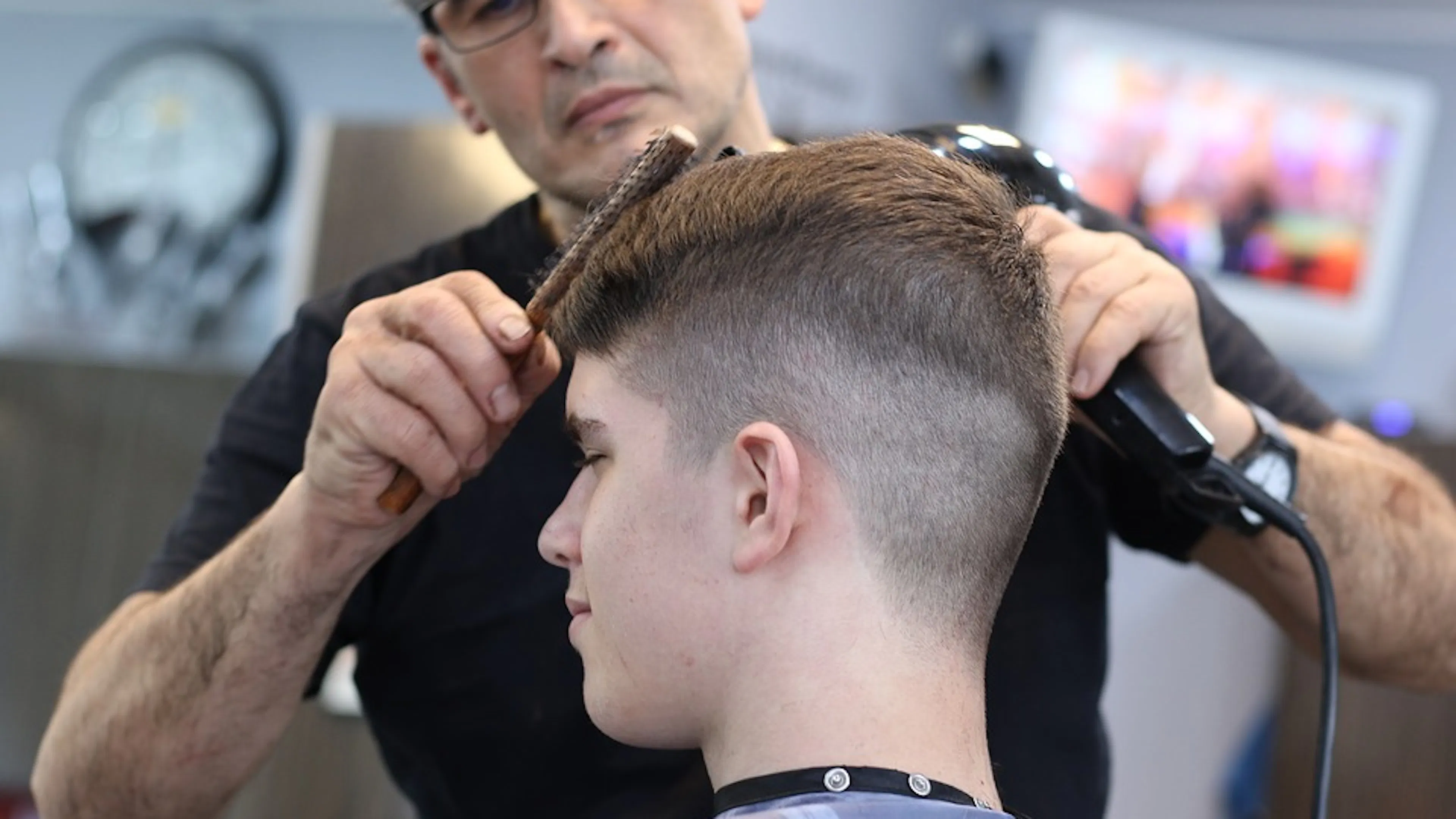 A barber using a comb and a clipper to trim a young man's hair in a military style cut
